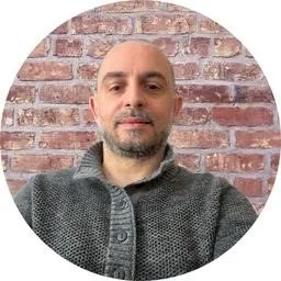 Portrait of a man with a beard and short hair in front of a brick wall, wearing a gray sweater.