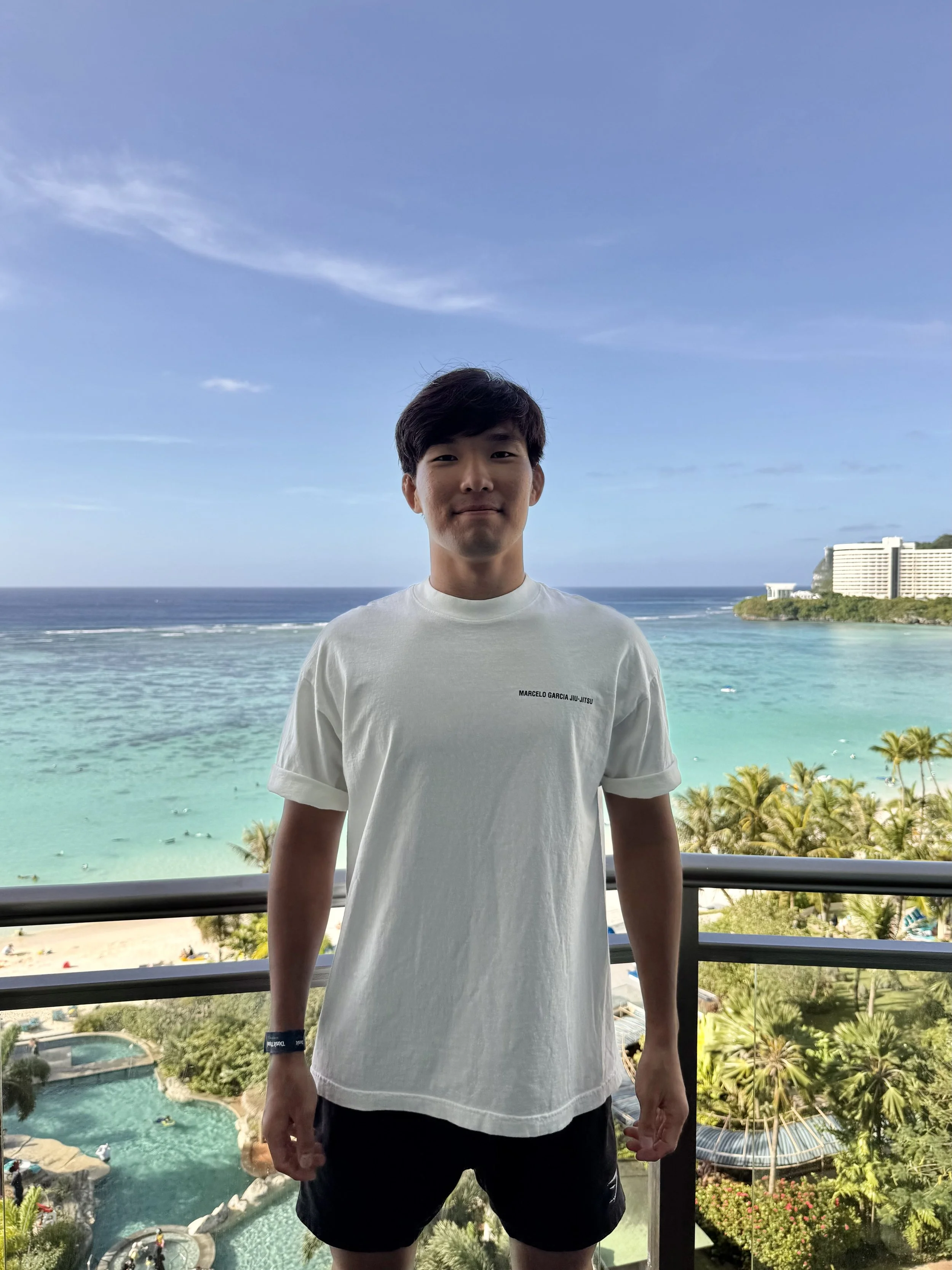 Jin Shin. Young man standing on a balcony overlooking a tropical beach with turquoise water, palm trees, and hotels in the background. Clipping MMA. Scale MMA.