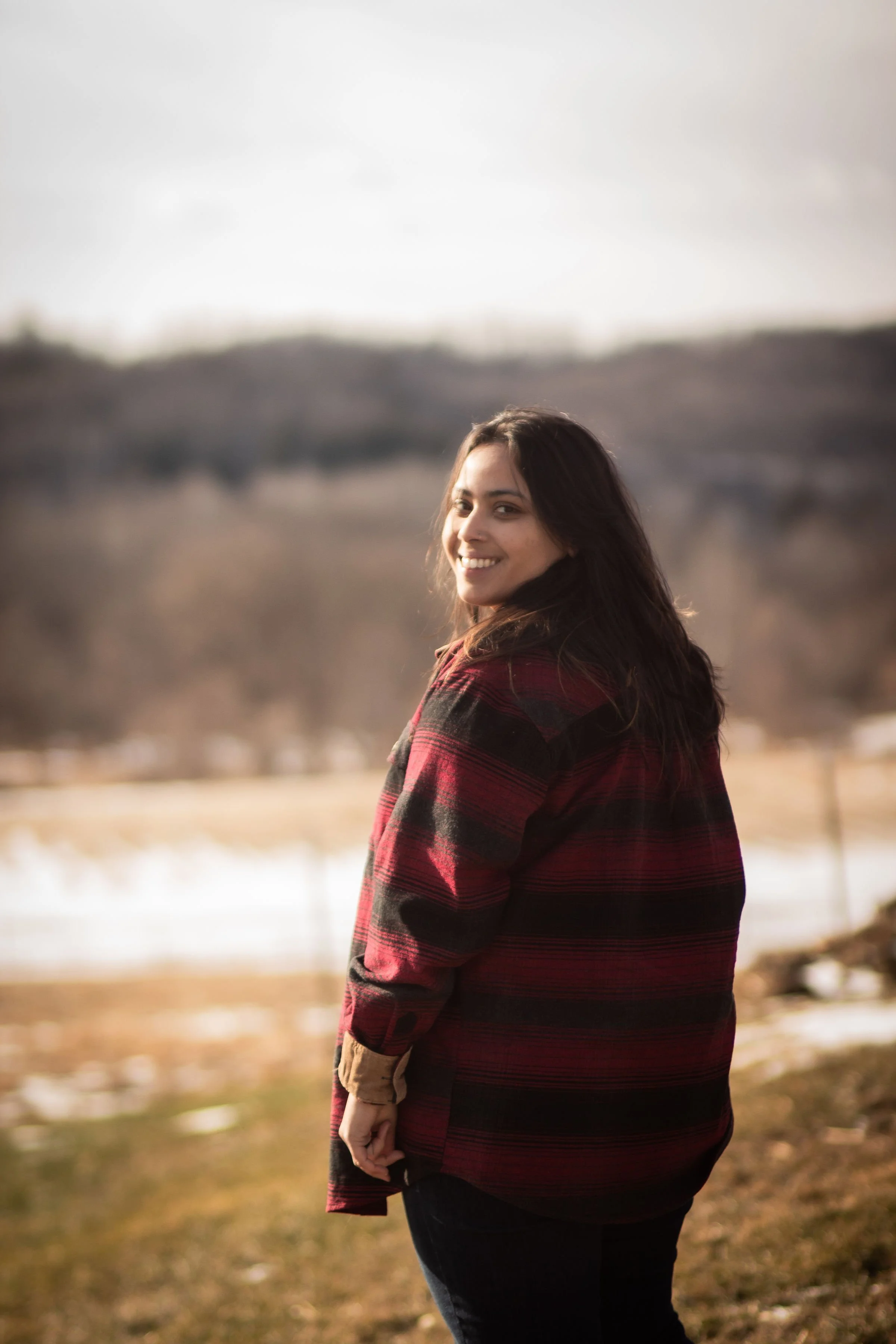 A woman with dark hair smiling and looking over her shoulder outdoors in a natural setting with hills and a body of water in the background, wearing a red and black plaid jacket.