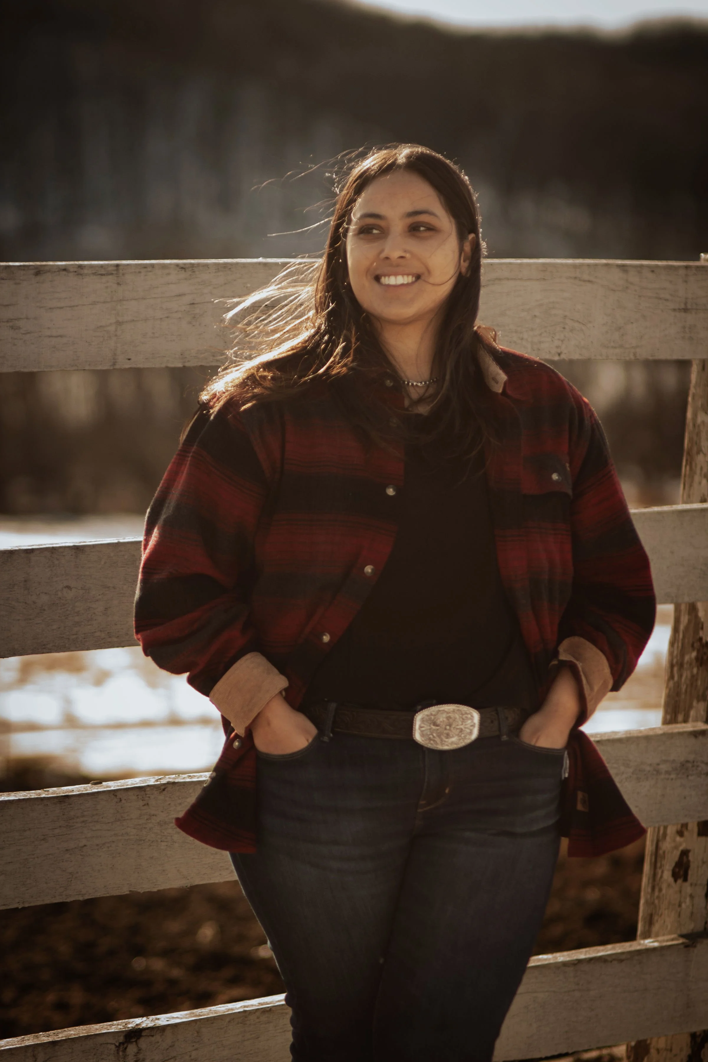 Young woman with dark hair smiling, wearing a black shirt, red plaid jacket, and jeans, leaning against a wooden fence outdoors with sunlight behind her.