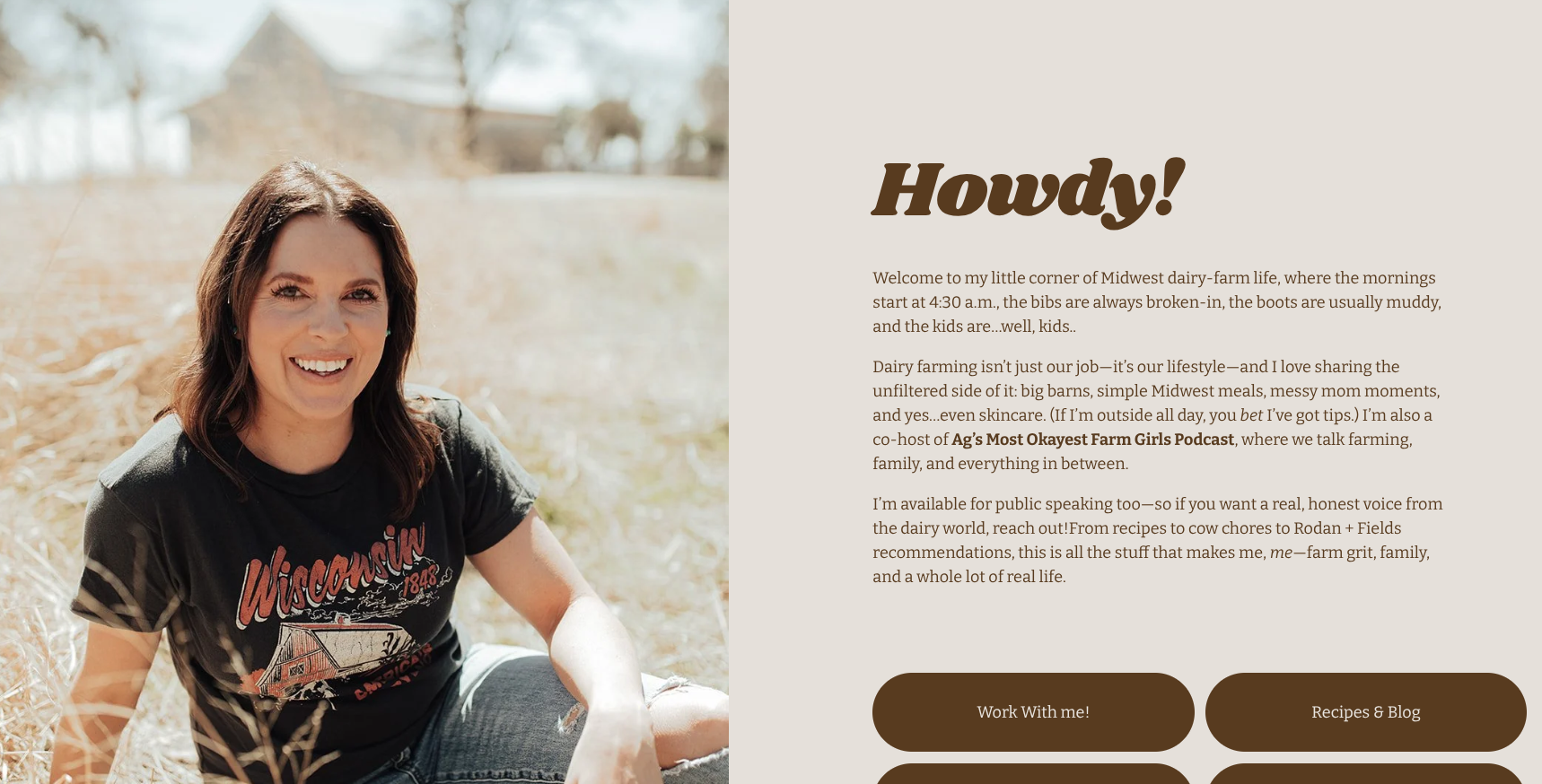 A smiling woman with dark brown hair, wearing a Wisconsin-themed t-shirt, sitting outdoors in a field of dry grass with trees and a building in the background.