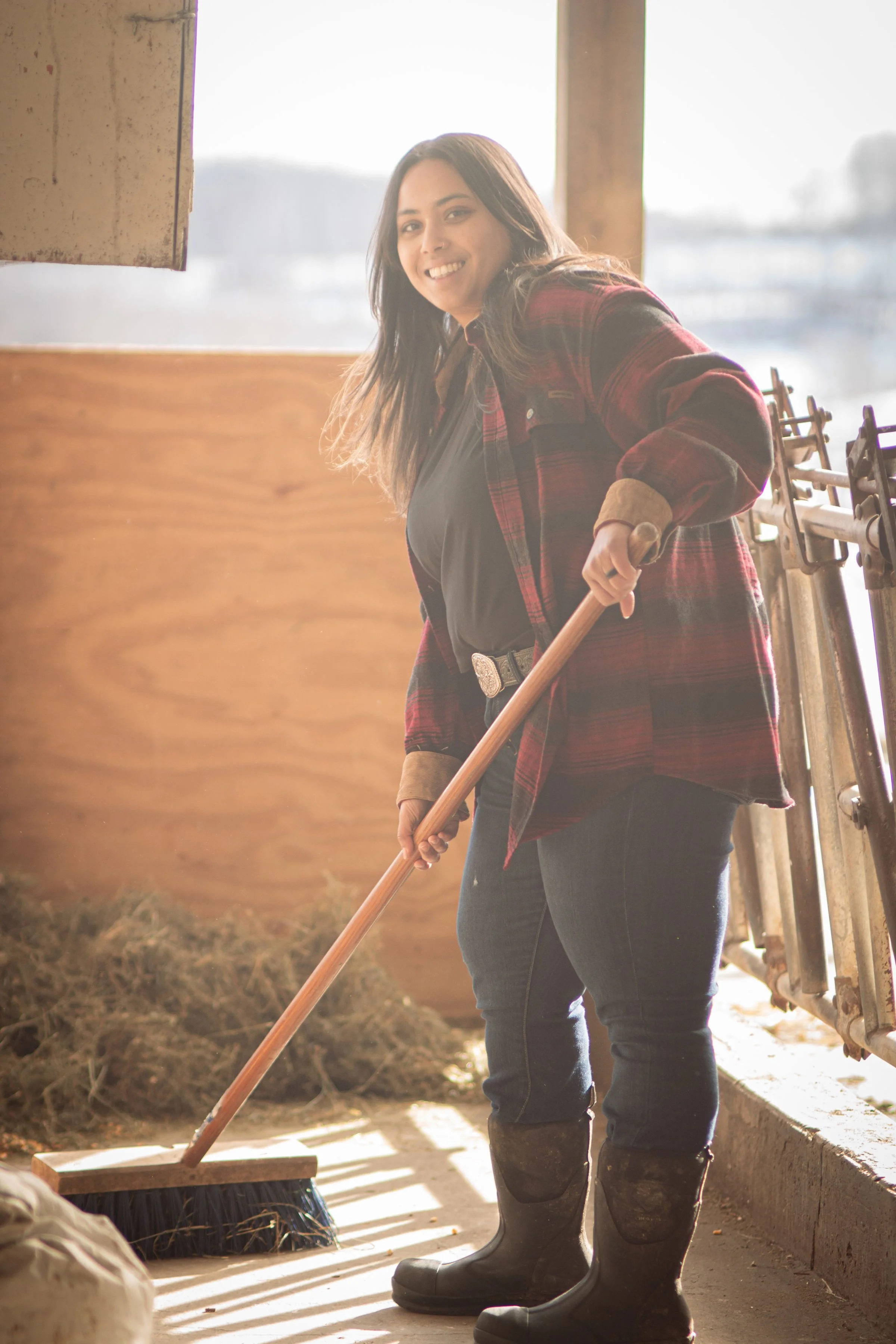 A woman with long dark hair wearing a red plaid jacket, black shirt, jeans, and rubber boots is sweeping the floor with a broom.