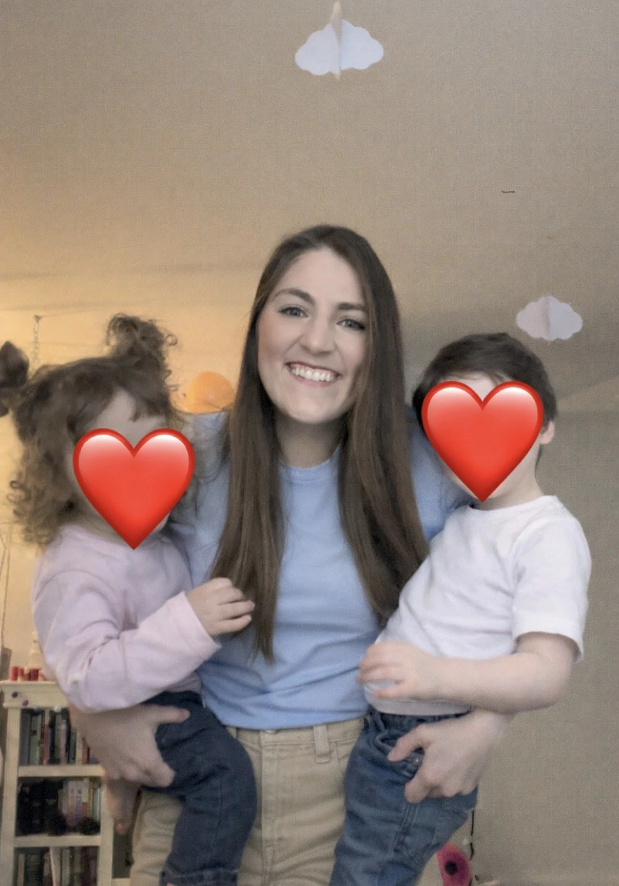 A smiling woman with long brown hair holding two children with heart emojis covering their faces, indoors with ceiling clouds decorations.