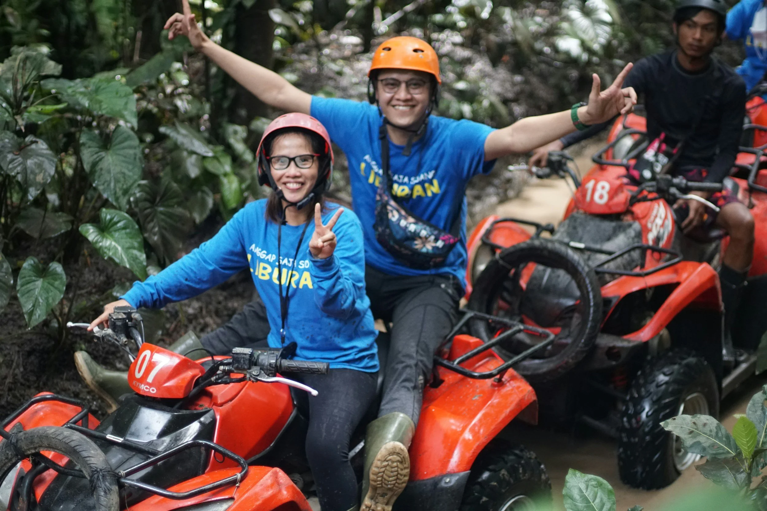 Two women and one man riding four-wheeler all-terrain vehicles through a forest. The women make peace signs and wear helmets, smiling and having fun.