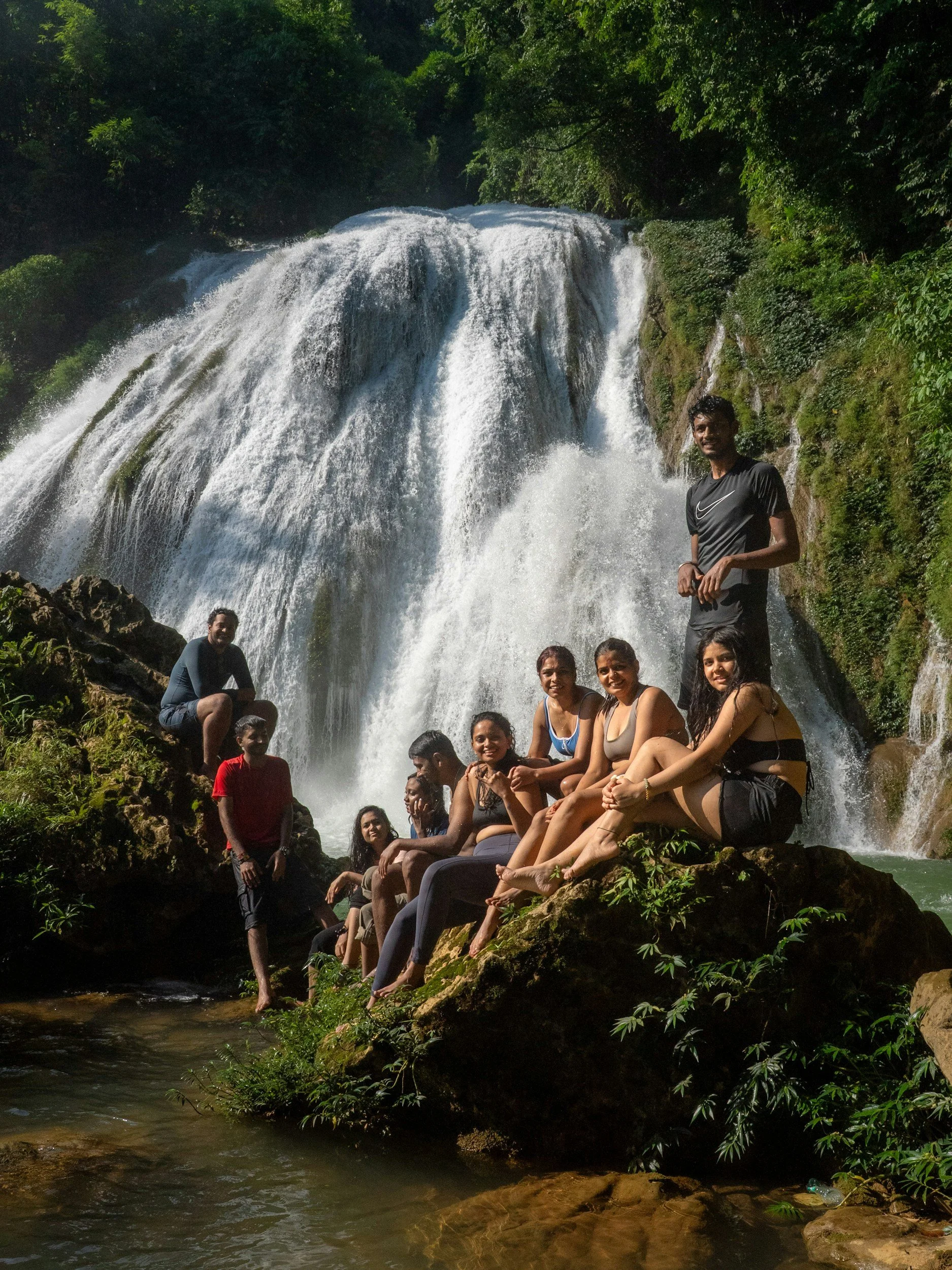 Group of people sitting on rocks in front of a waterfall in a lush green forest