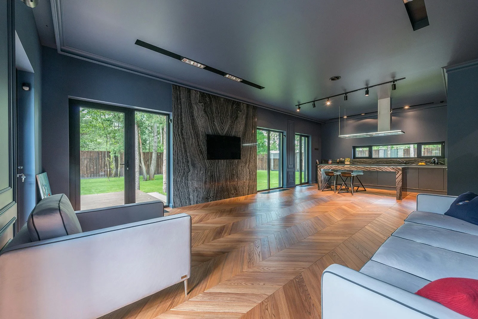 Sala moderna con paredes grises, ventanas grandes con vista al jardín, piso de madera en patrón de espiga, sofá gris claro y sillón blanco con cojines, pared decorativa de piedra, televisión montada, cocina en el fondo con barra y sillas, luz artificial en el techo.