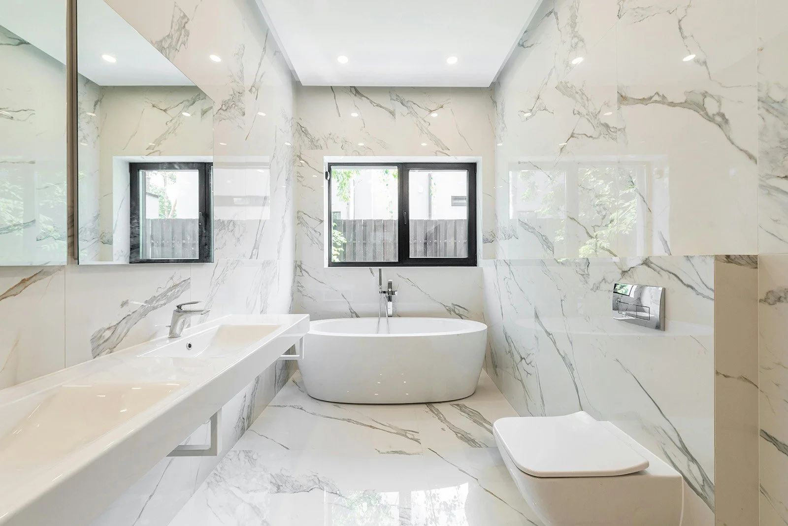 Un baño moderno con paredes y piso de mármol blanco con vetas grises, una bañera independiente, un lavabo con espejo, ventanas grandes y un inodoro en color blanco.
