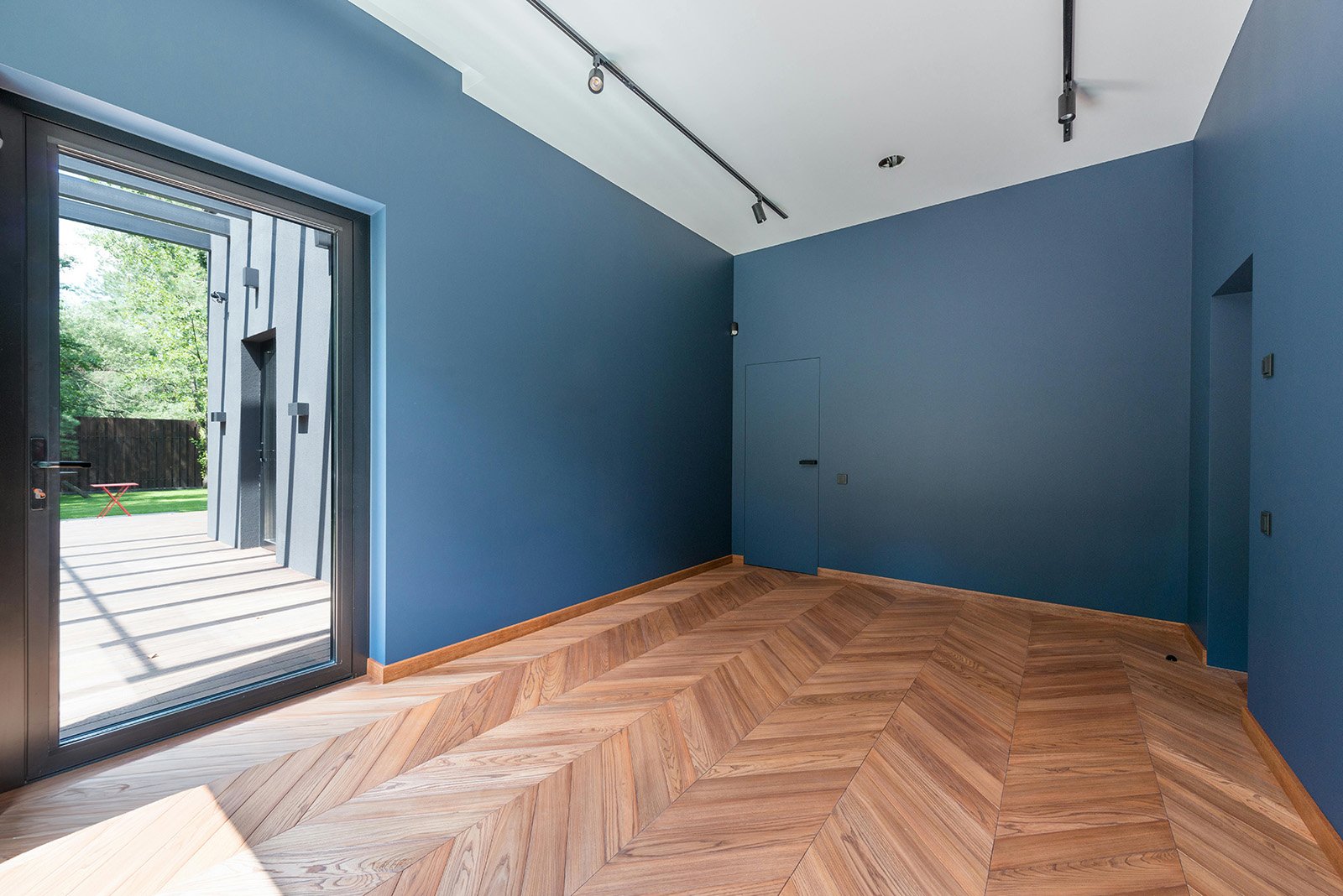  habitación vacía con paredes azules, piso de madera en patrón diagonal, puerta de cristal que da a un patio con césped y árboles, y techo blanco con iluminación de rieles