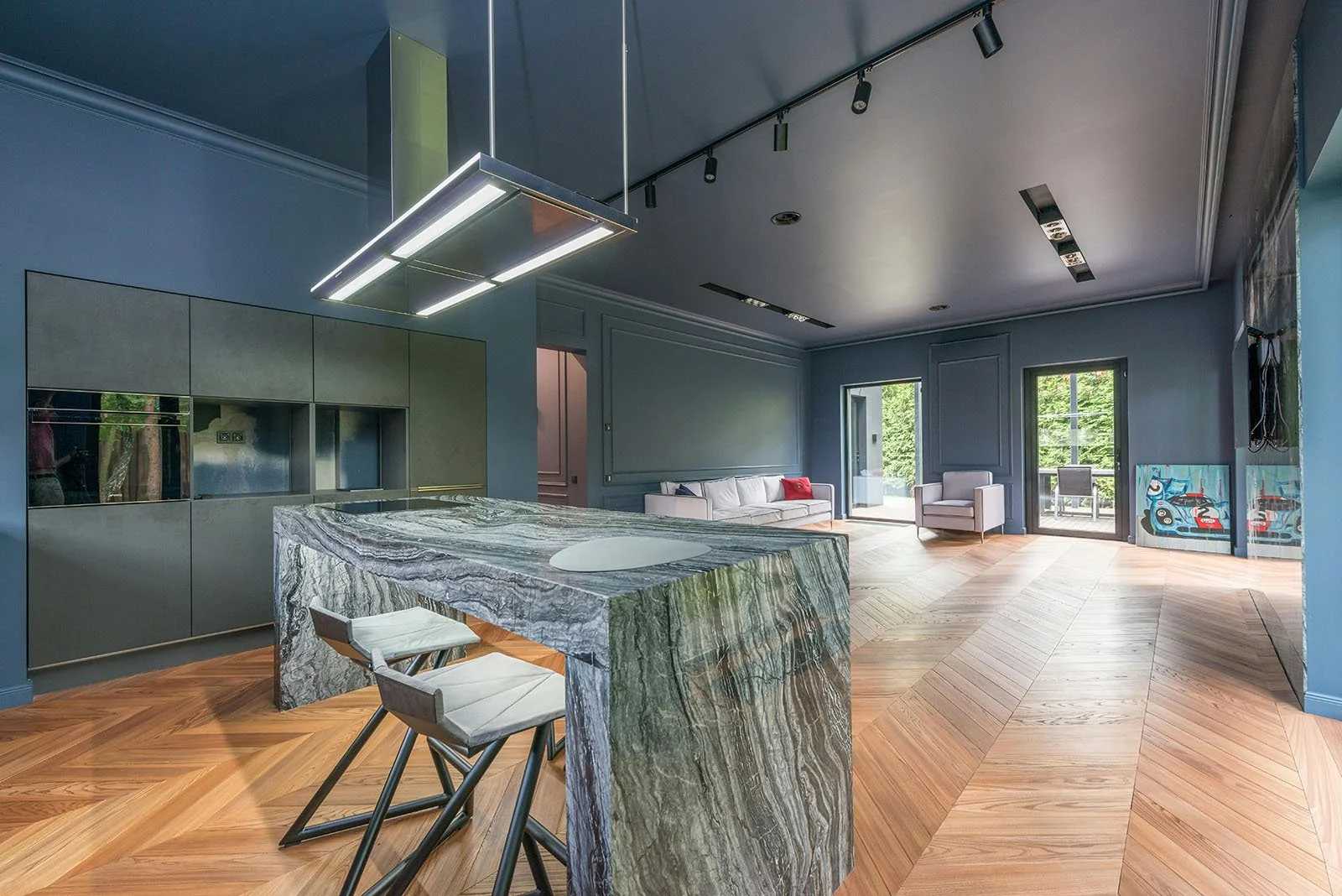 Interior de una sala moderna con paredes azul oscuro, piso de madera y muebles blancos, incluyendo un sofá y sillones, con ventanas que dejan ver árboles verdes y arte en el suelo.