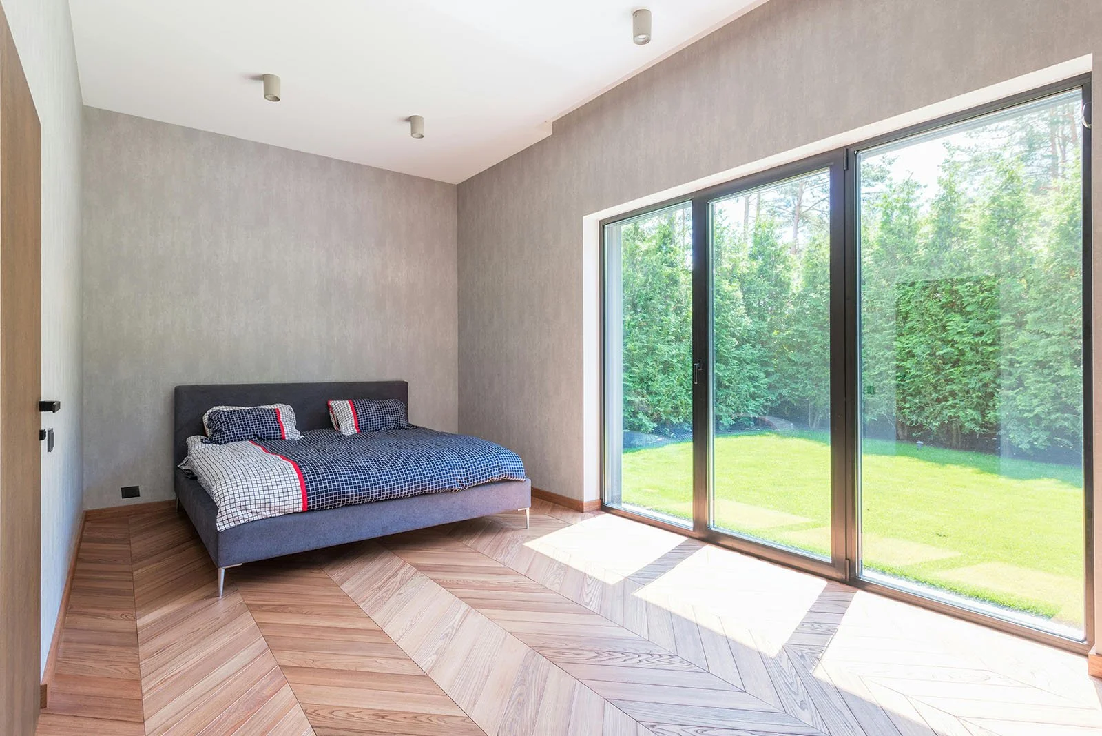 Habitación moderna con cama de colchón gris y ropa de cama a cuadros en azul, blanco y rojo, piso de madera en patrón diagonal y grandes ventanales que permiten vista a un jardín con árboles verdes.