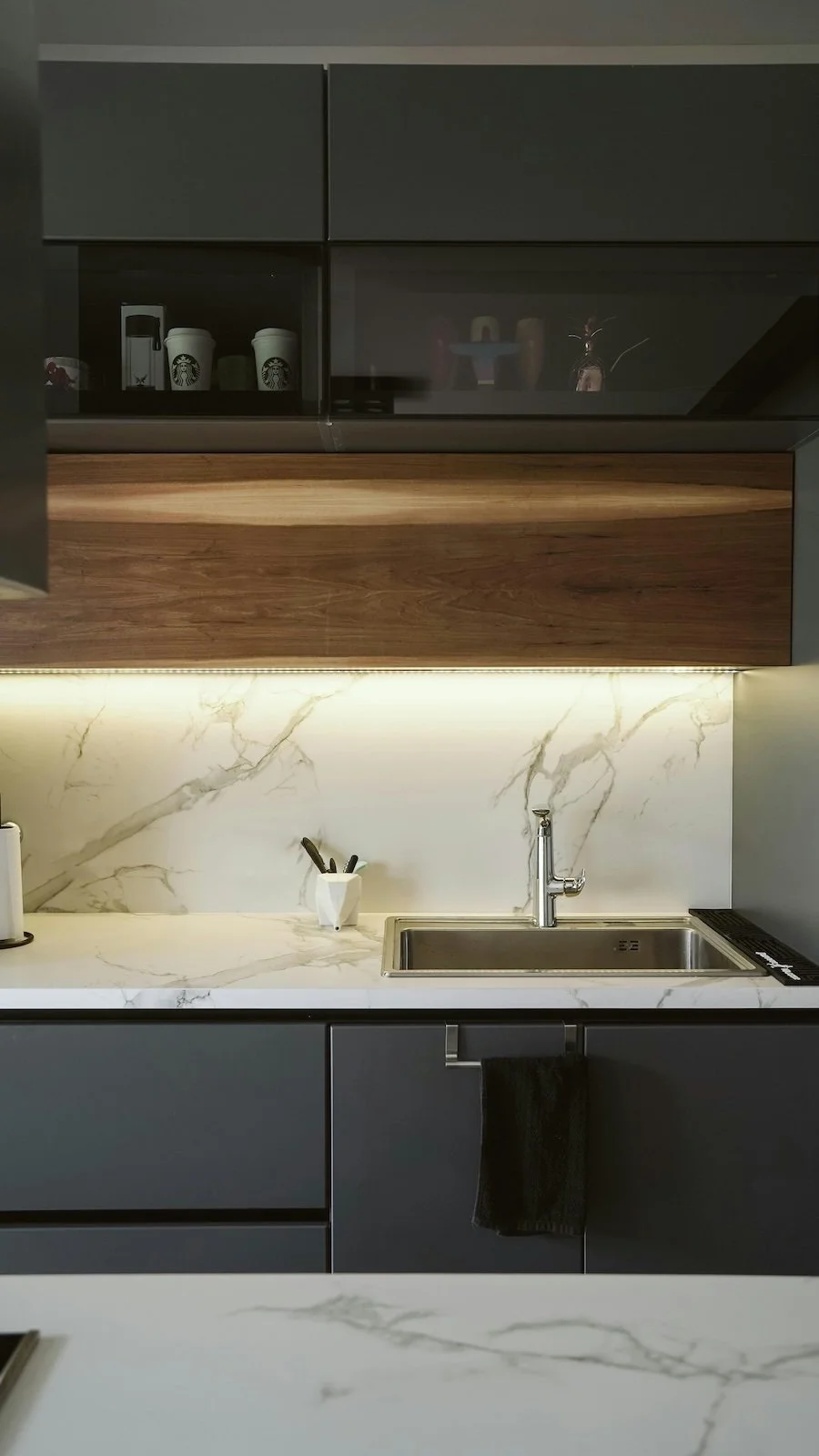 Cocina moderna con encimera de mármol blanco, armarios grises y una vitrina con tazas Starbucks y electrodomésticos en la parte superior.