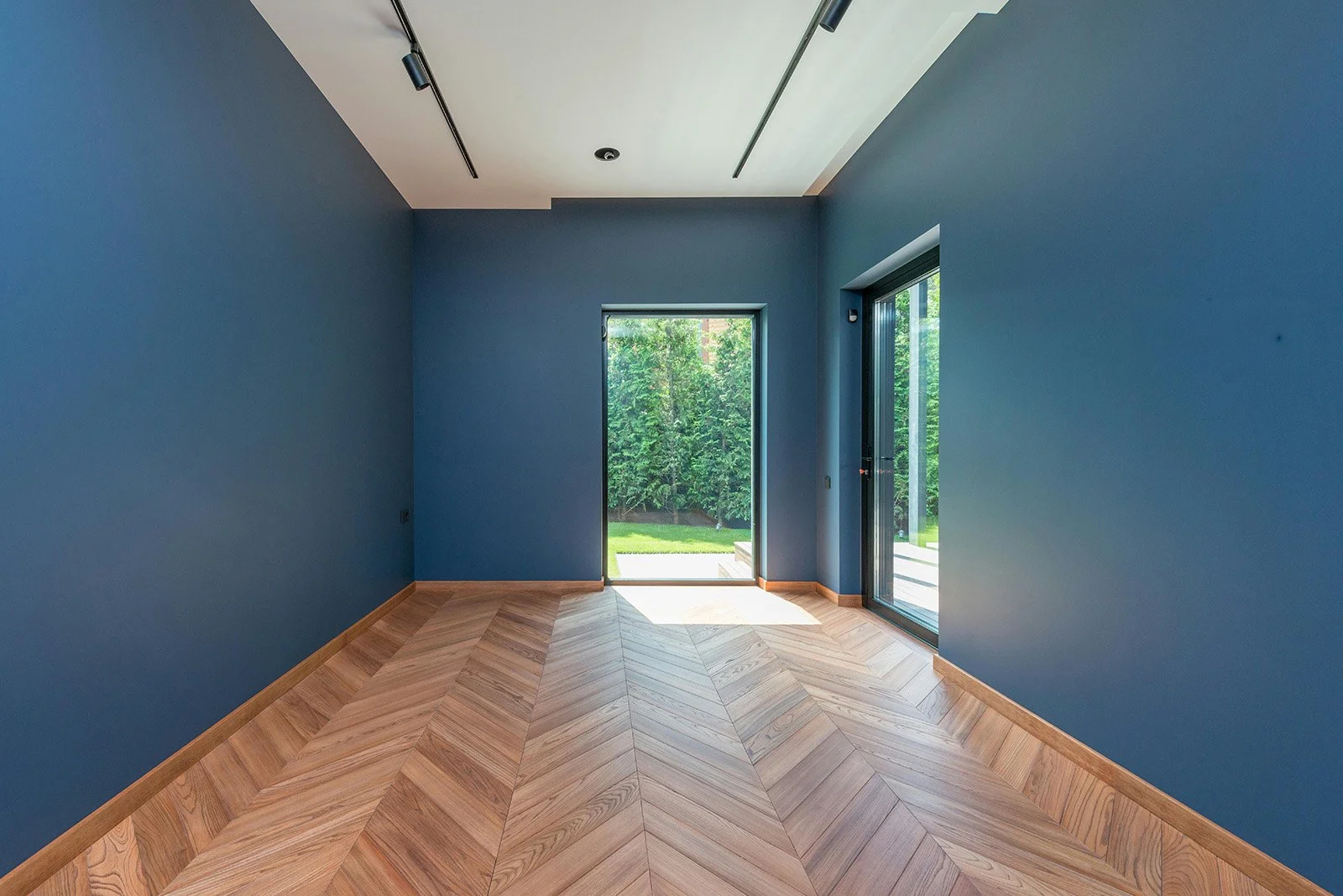 Sala vacía con paredes azules, suelo de madera en patrón de espiga, puertas de vidrio que dan al jardín exterior con césped y árboles.