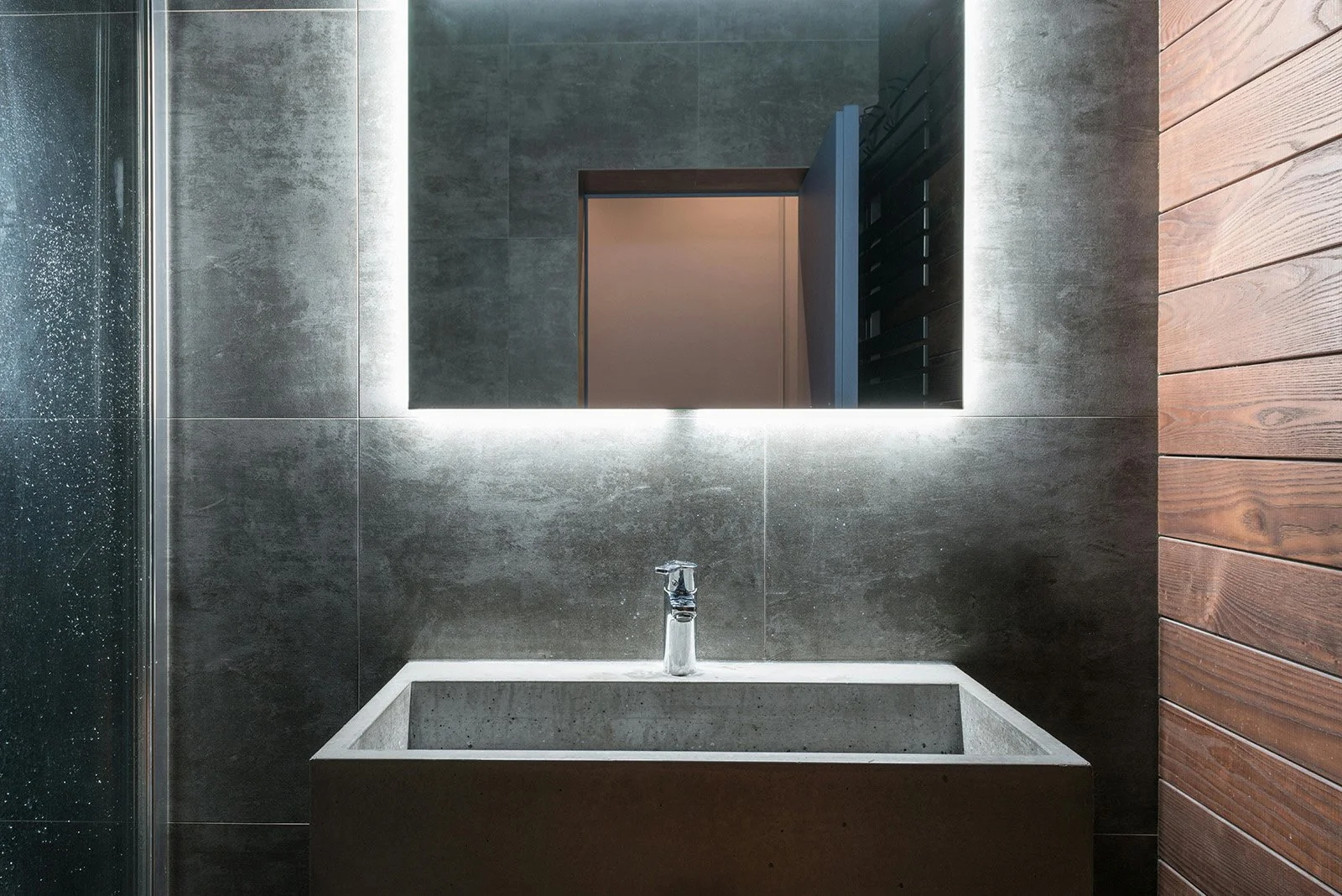Lavabo de concreto debajo de un espejo con iluminación LED frontal en un baño moderno con paredes de cerámica gris y pared de madera a la derecha.