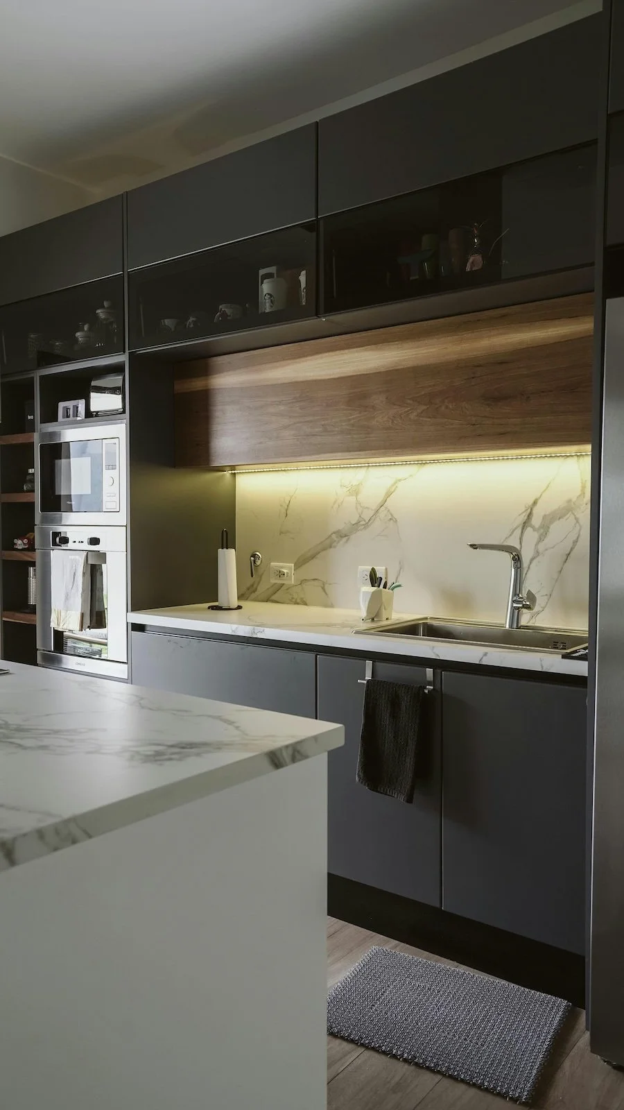 Cocina moderna con gabinetes grises, encimera de mármol, backsplash de mármol, y estantes de madera, con electrodomésticos integrados y luces debajo de los gabinetes.