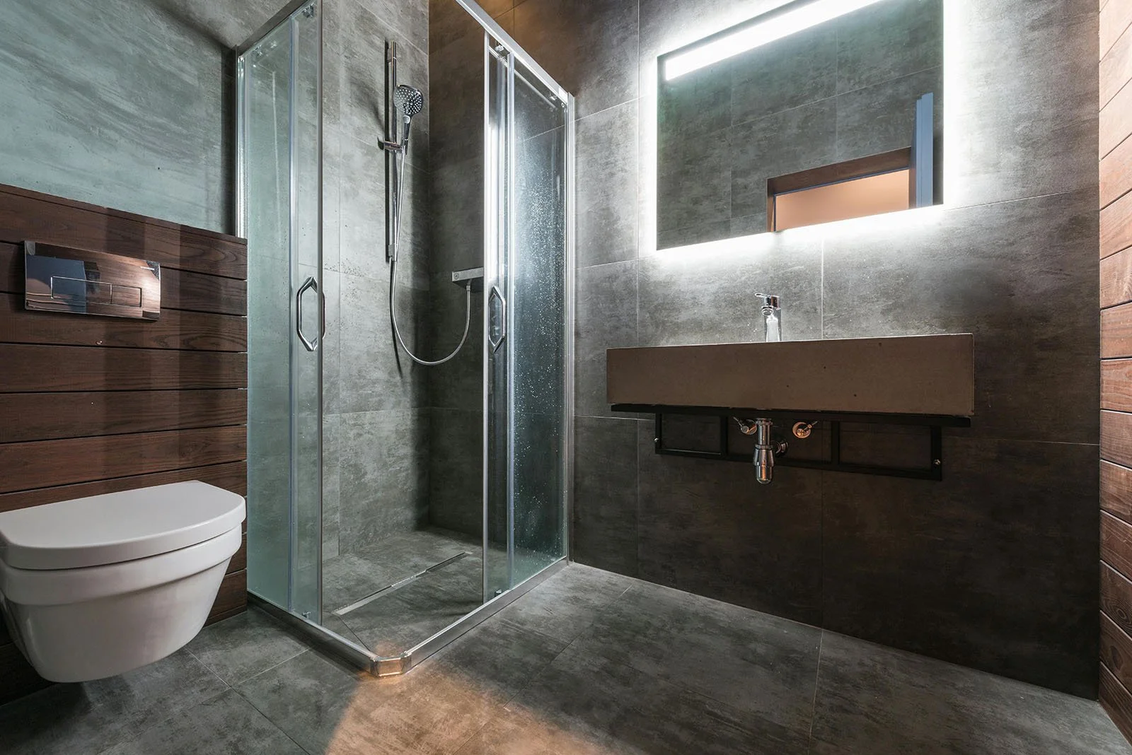 Baño moderno con ducha, inodoro y lavabo con espejo con luz LED en paredes de piedra gris oscuro y detalles de madera.