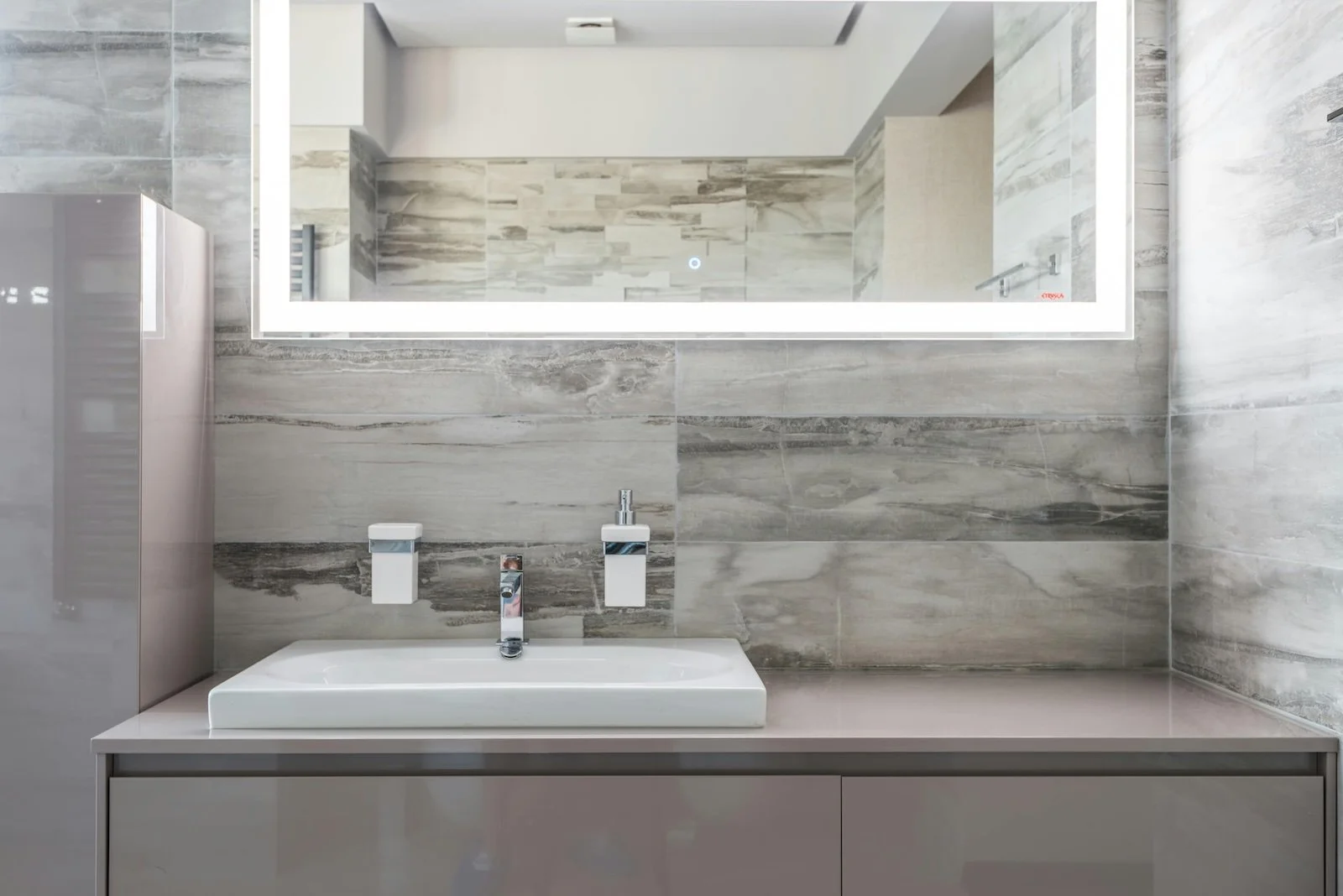 Lavabo en un baño con pared de baldosas de tonos grises y un gran espejo con luz LED en la parte superior.