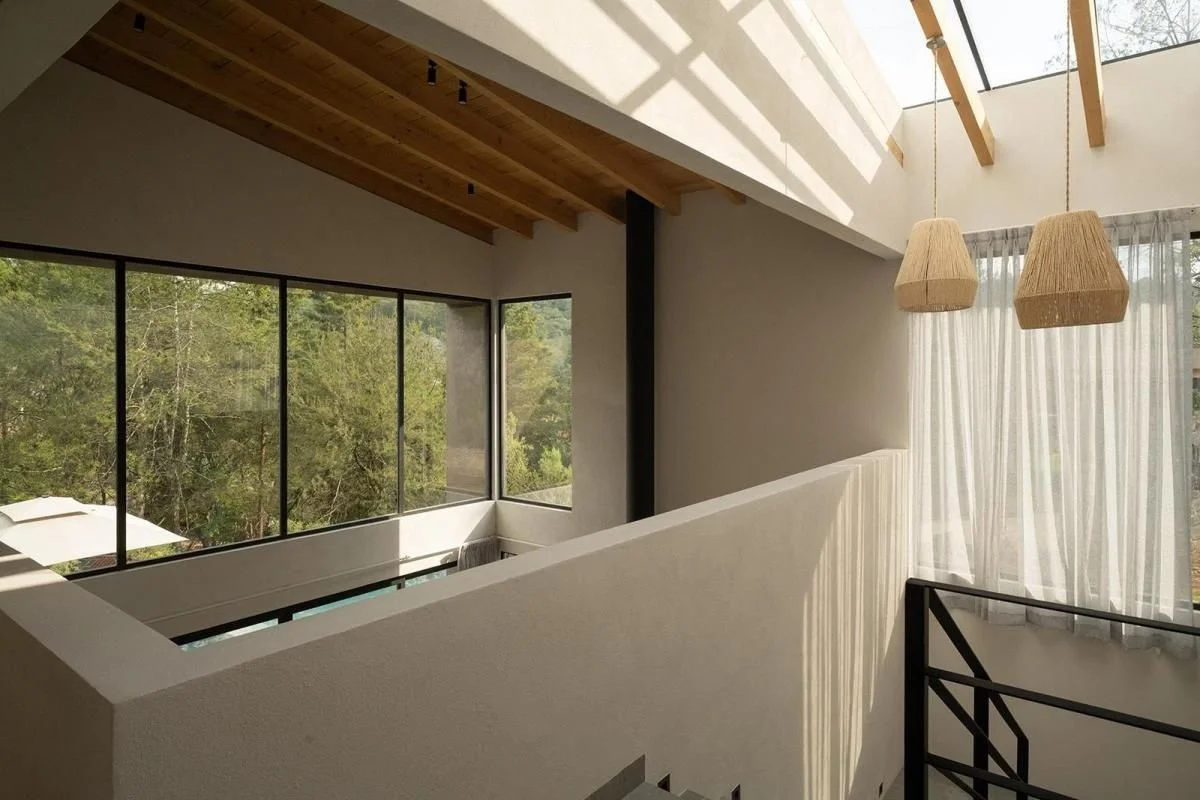 Interior de una casa moderna con ventanas grandes, paredes blancas, techo de madera expuesta, cortinas blancas, lámparas de fibra natural y un techo con claraboya que deja entrar luz natural.