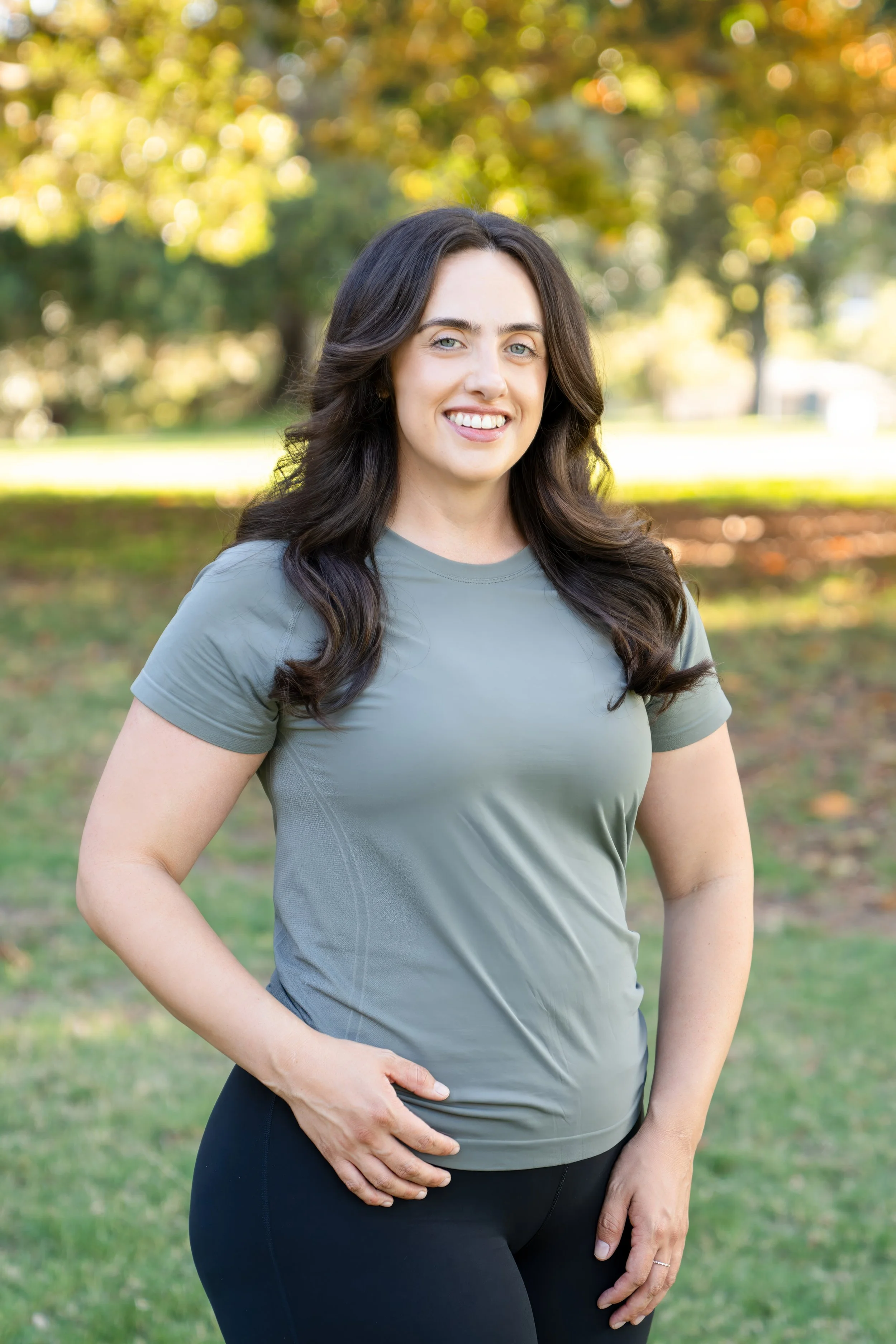 A smiling woman with long dark hair stands outdoors in a park on a sunny day, wearing a gray sports t-shirt and black leggings.