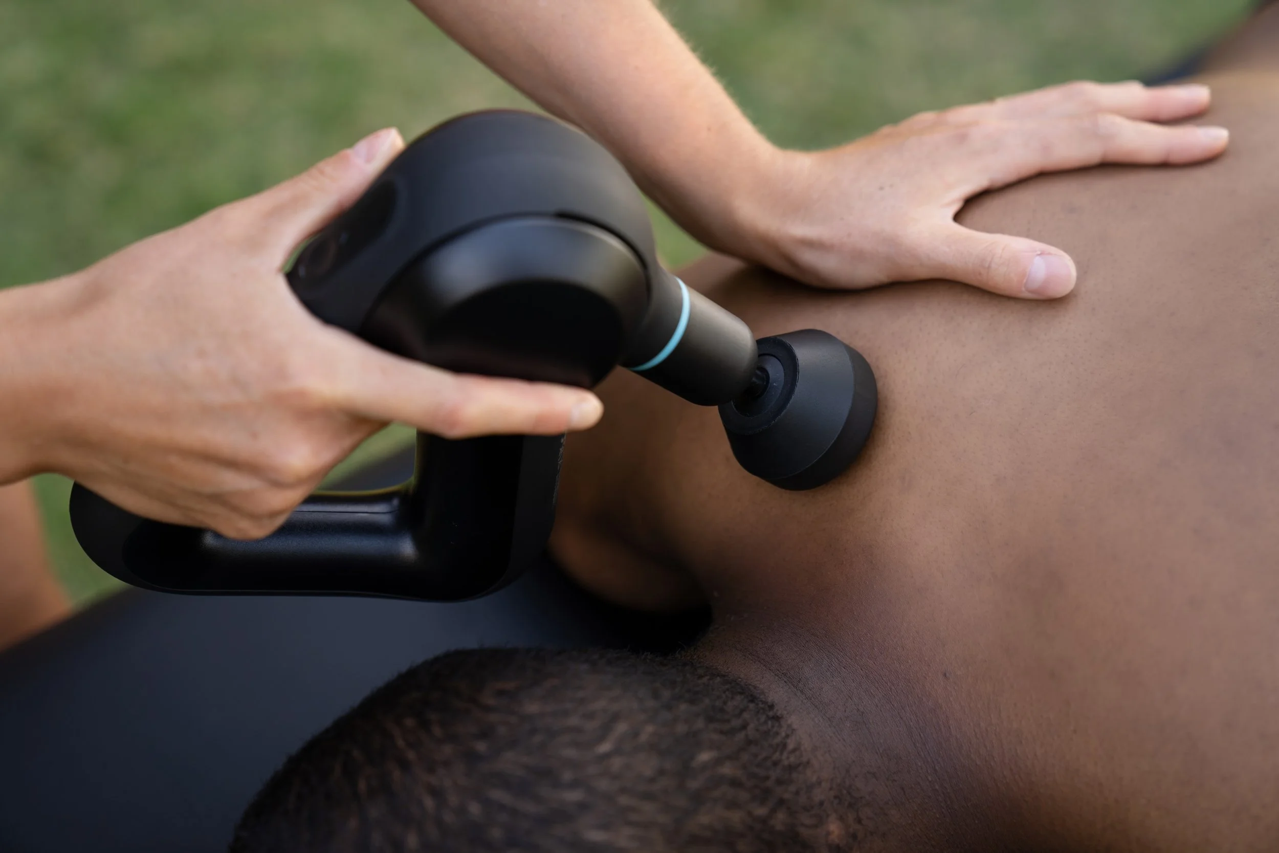 A person receives a massage using a black massage gun on their back outdoors.