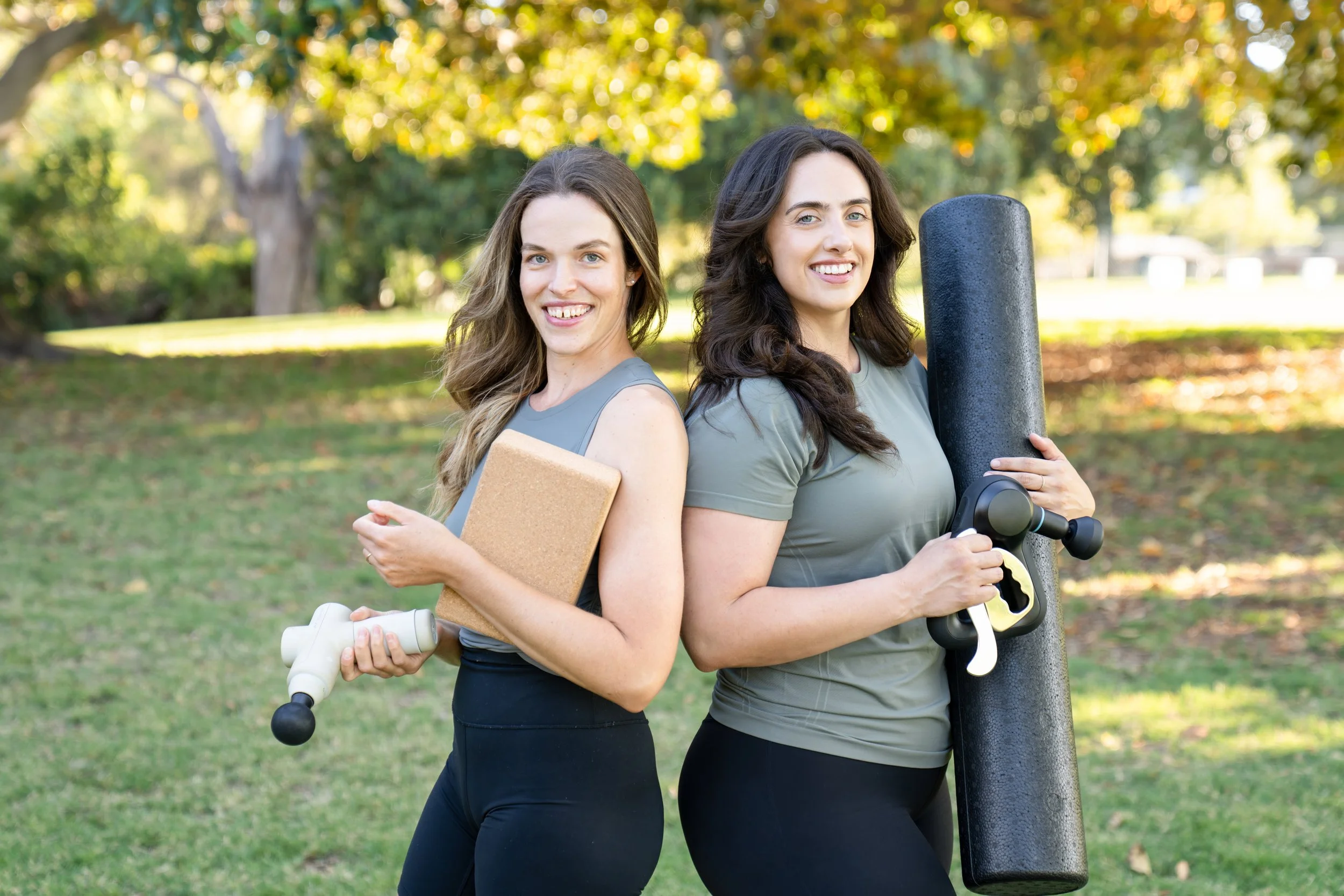 Two women stand back-to-back outdoors, smiling and holding fitness equipment, with trees and sunlight in the background.
