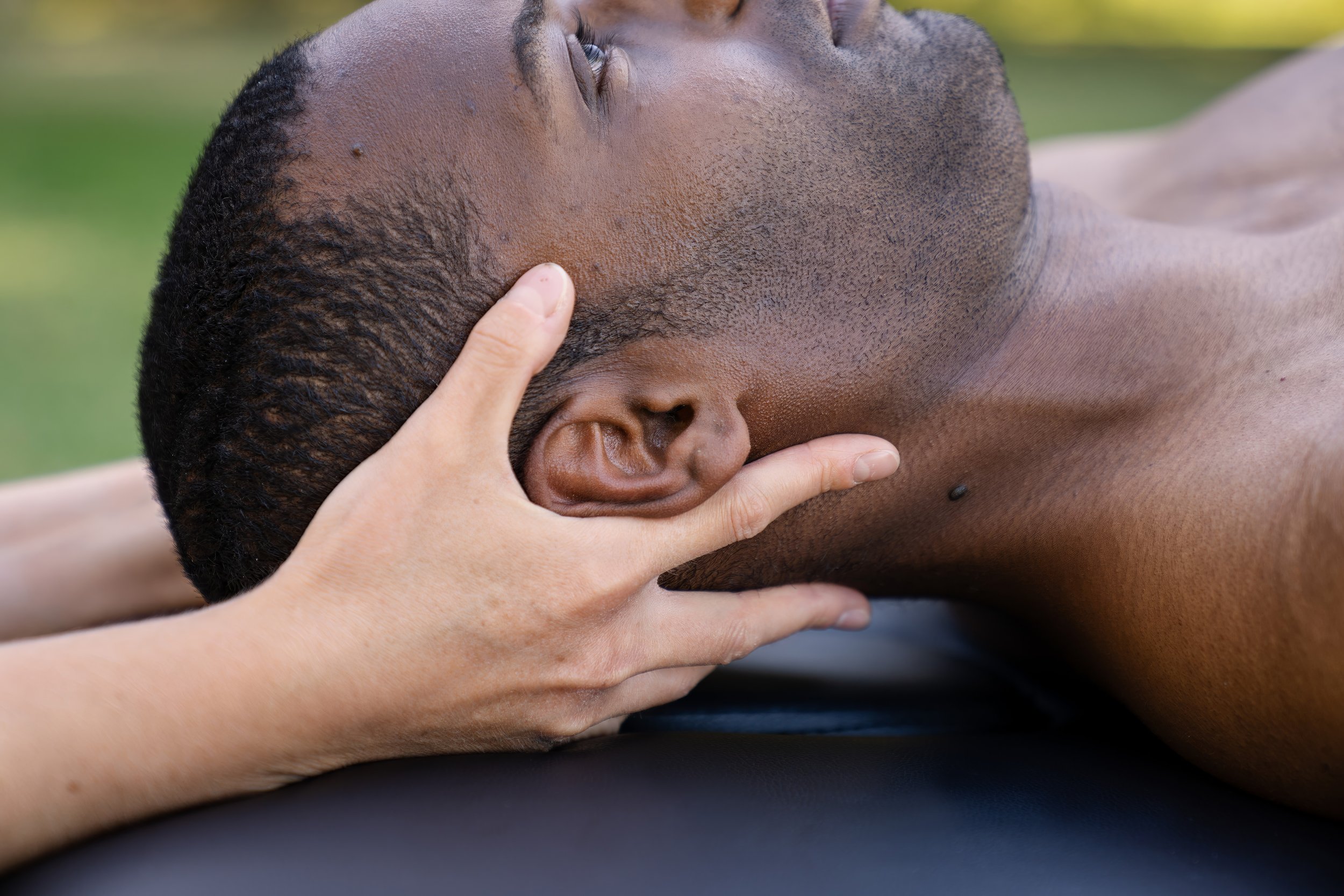 A person with dark skin lying on their back outdoors with a hand on their head and neck, eyes closed, and short dark hair.