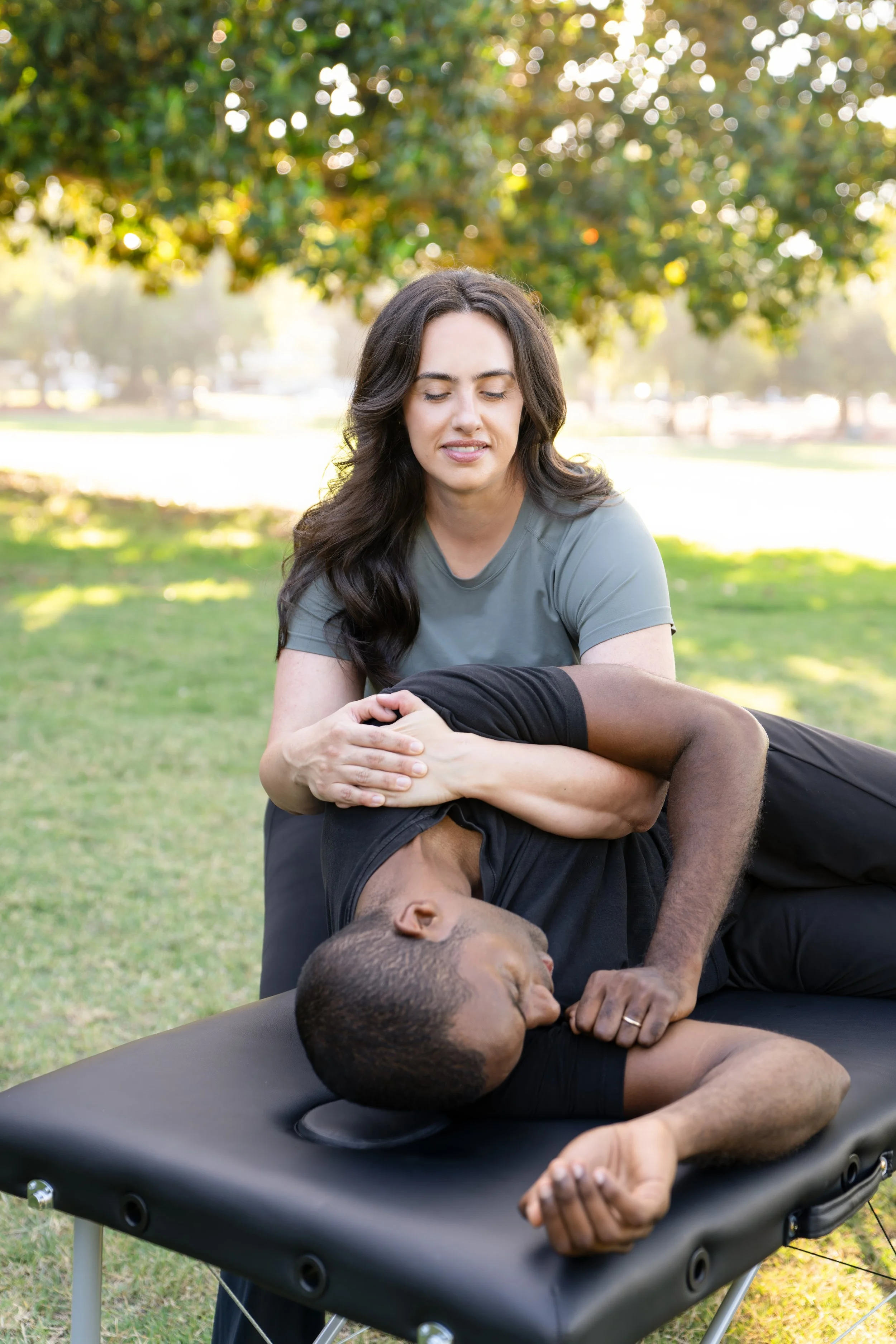 A woman practicing acro yoga outdoors on a trampoline, with a man lying on a massage table beneath her, holding his hand and smiling. The scene is set in a park with trees and sunlight in the background.