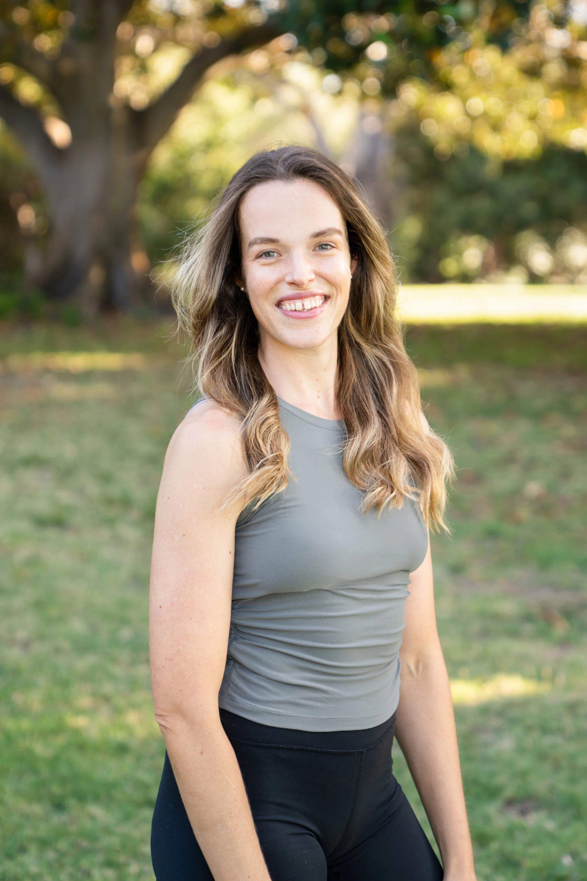 A woman with long, wavy hair and light skin smiling outdoors in a park with green grass and trees in the background, wearing a gray athletic tank top and black workout pants.