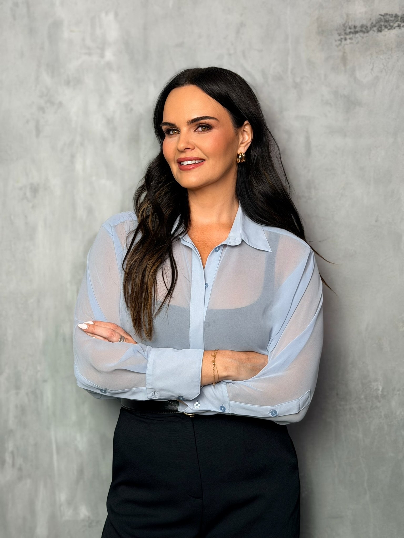 A woman with dark hair wearing a light blue blouse and black pants, standing with her arms crossed against a concrete wall background.
