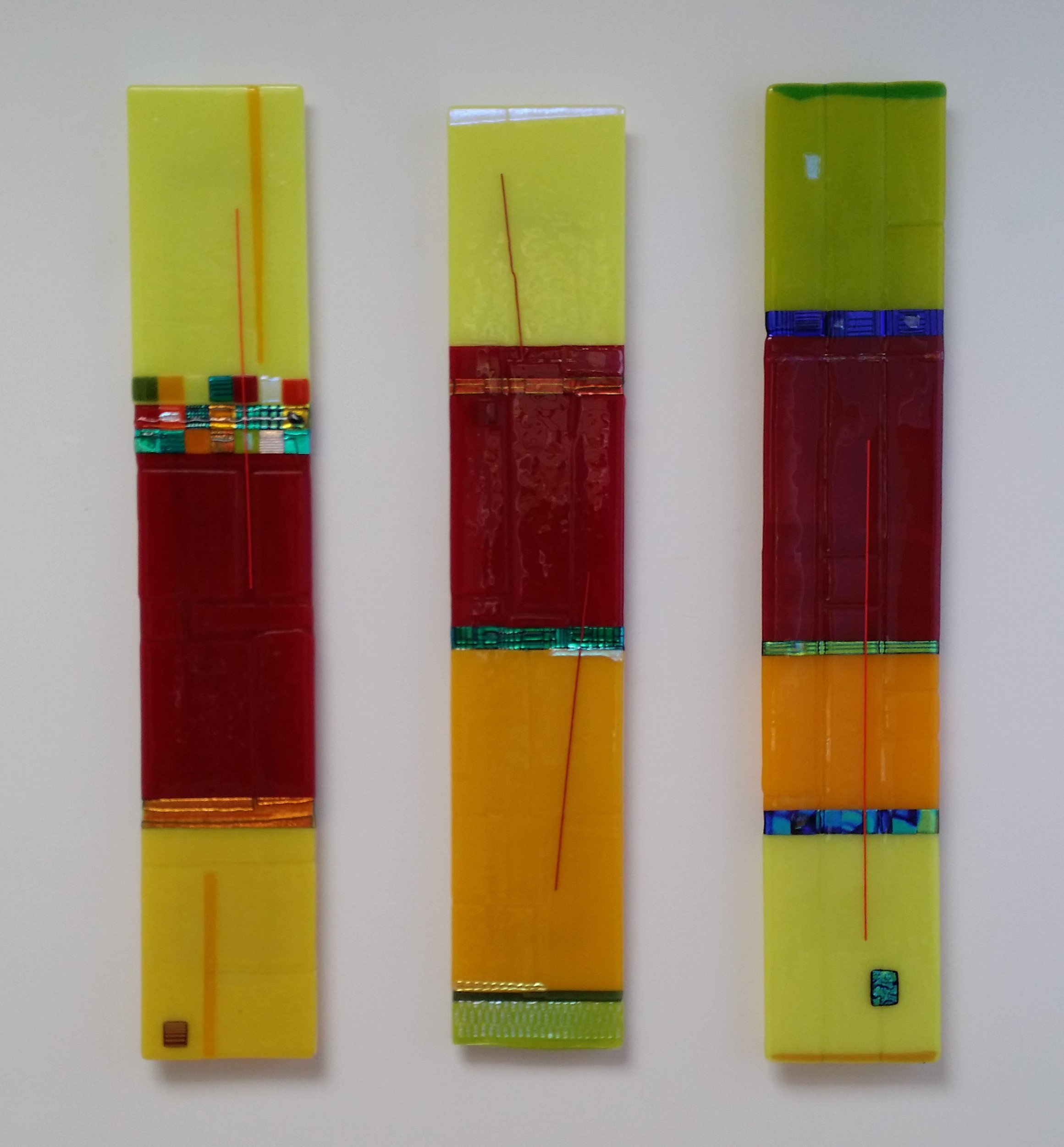 Three vertical colorful glass art pieces with rectangular shapes composed of yellow, red, green, blue, and orange sections, mounted on a light gray wall.