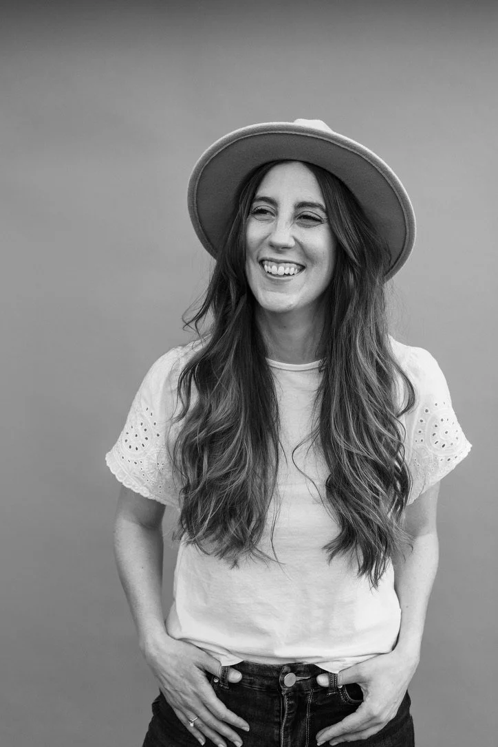 A woman with long wavy hair, wearing a hat and a short-sleeved eyelet blouse, smiling and posing with hands in her pockets.