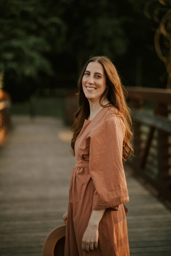A woman with long, wavy brown hair smiling and standing on a wooden bridge outdoors during sunset, wearing a rust-colored dress and holding a hat.