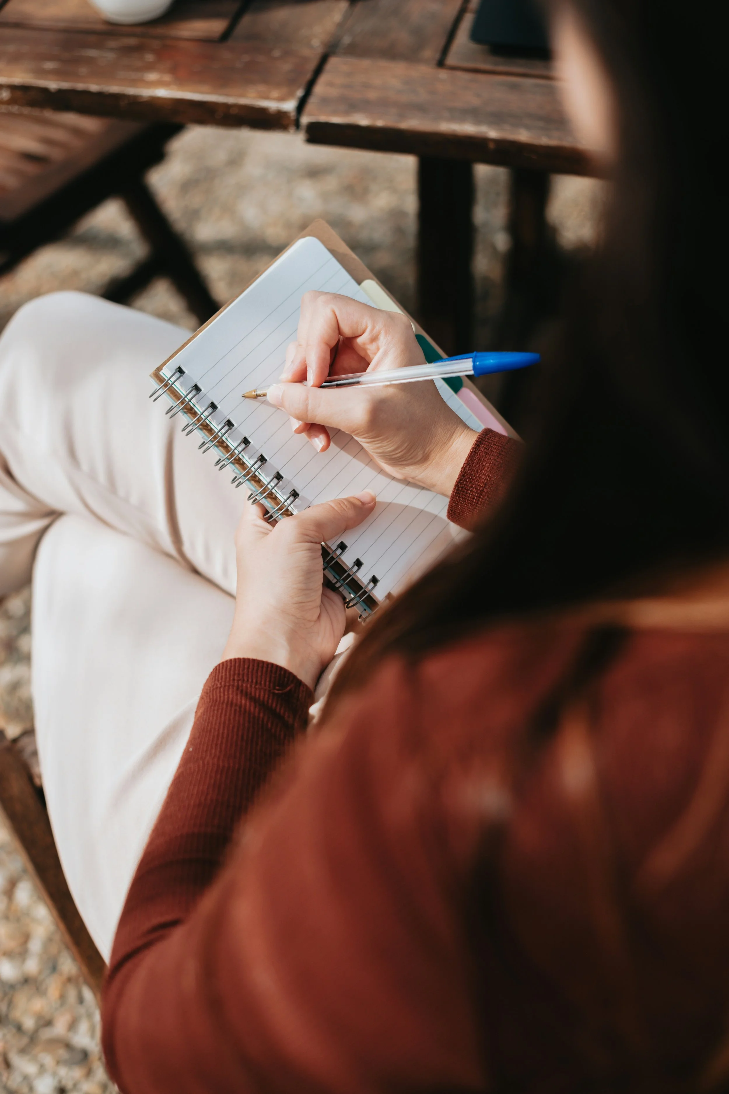 A person wearing a brown long-sleeve shirt writing in a spiral notebook with lined pages using a blue capped pen, seated outdoors at a wooden table.