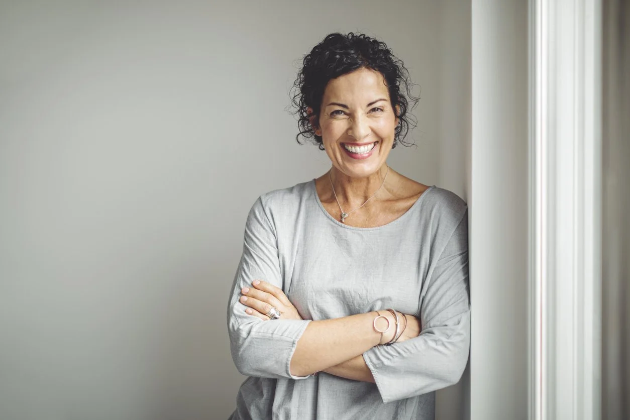 A smiling woman with curly dark hair standing by a window with arms crossed.