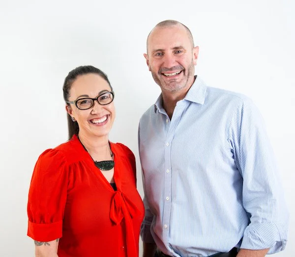 A smiling woman wearing glasses and a red blouse standing next to a smiling man in a light blue striped shirt against a white background.