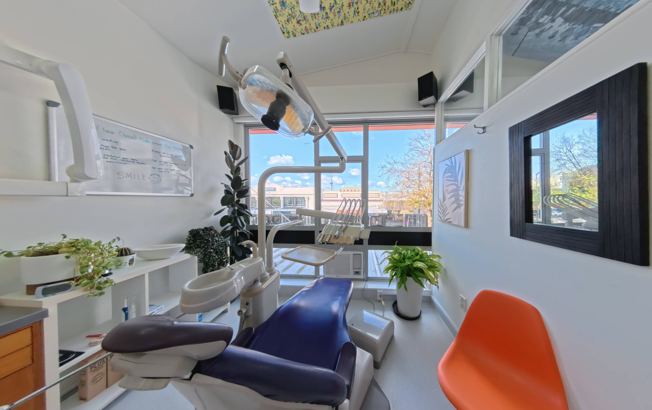 Interior view of a dental office with dental chair, equipment, plants, window view of buildings and trees, and decorative framed art on walls.