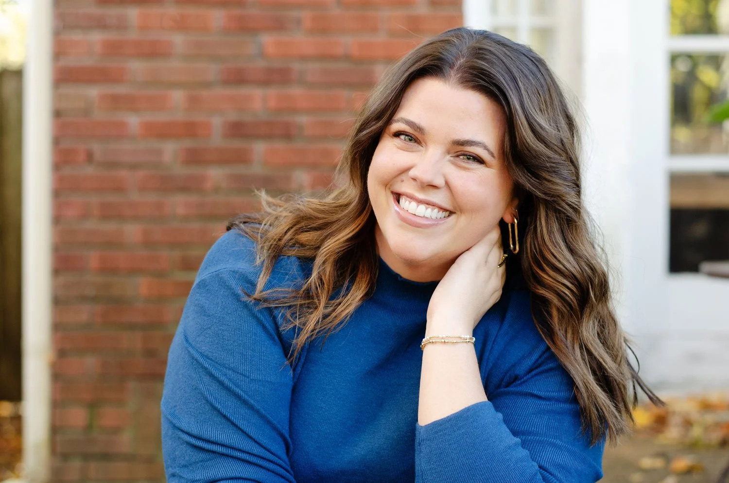 A smiling woman with long wavy brown hair, wearing a blue sweater, and gold hoop earrings, outdoors in front of a brick wall and a house with white siding, during autumn.