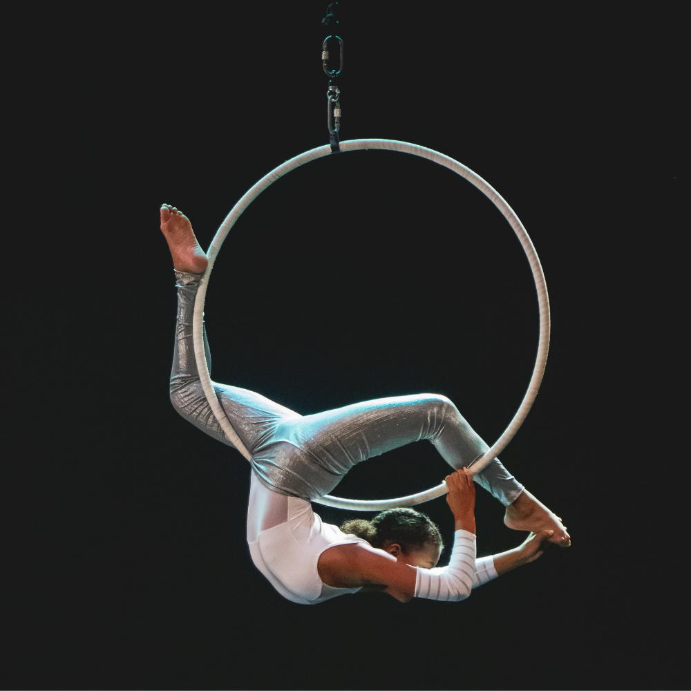 A female aerialist in silver and white costume performing on a suspended aerial hoop against a dark background.