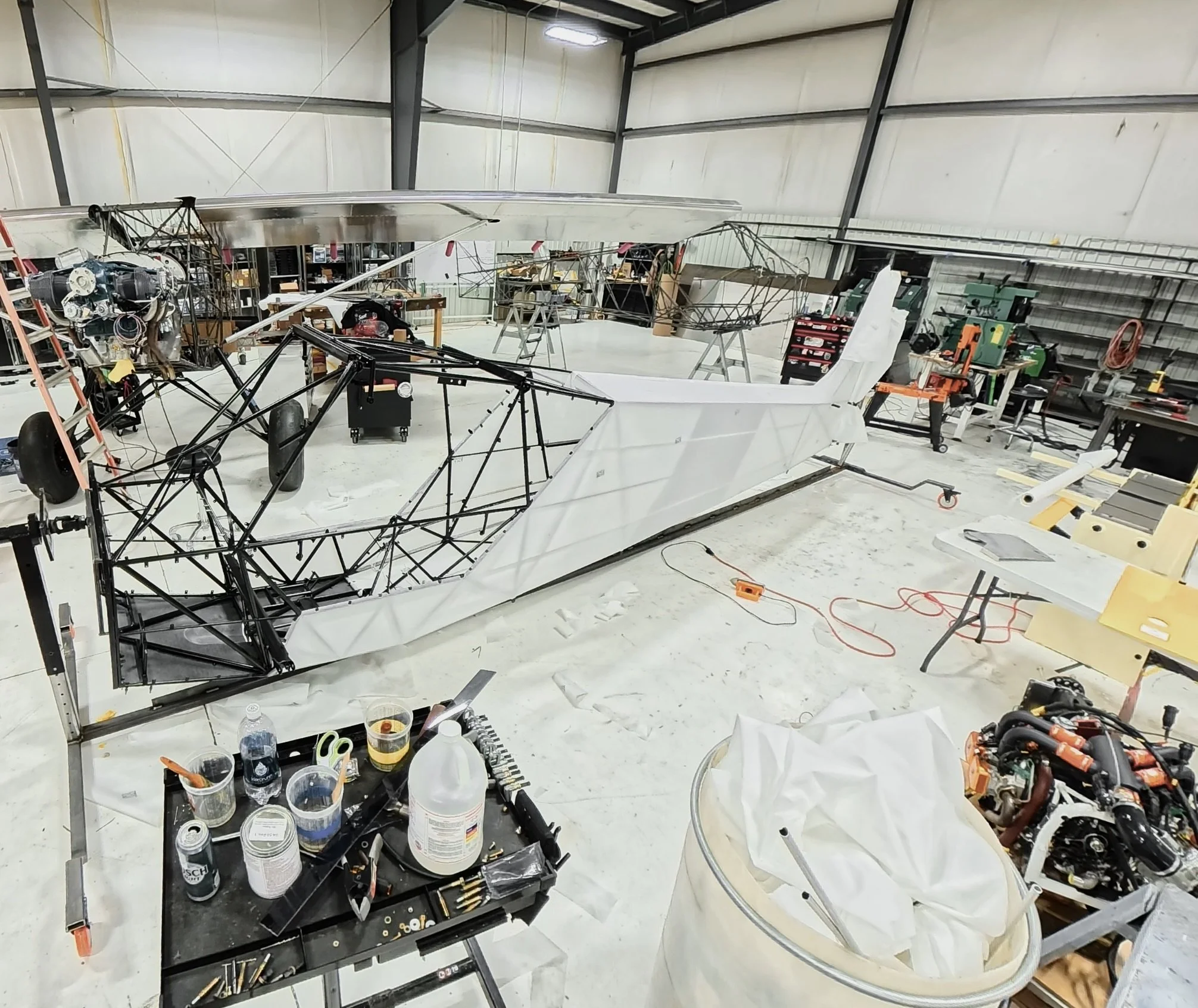 An indoor workshop space with a partially assembled small aircraft or glider, surrounded by tools and equipment, including a workbench, power tools, and parts.