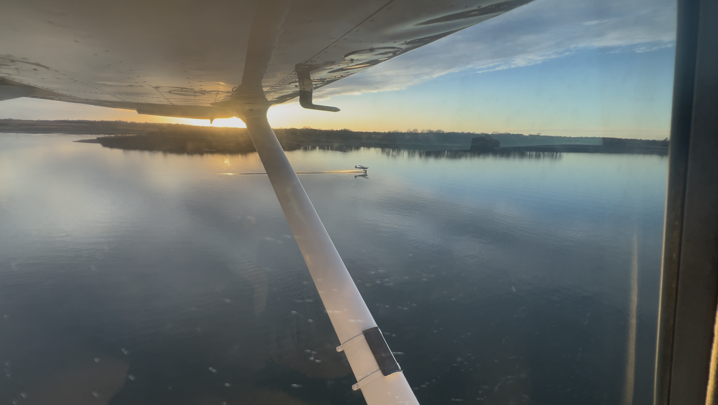 View from an airplane window showing the underside of the wing, a body of water reflecting the sky, a boat leaving a wake, and a distant land at sunset or sunrise.