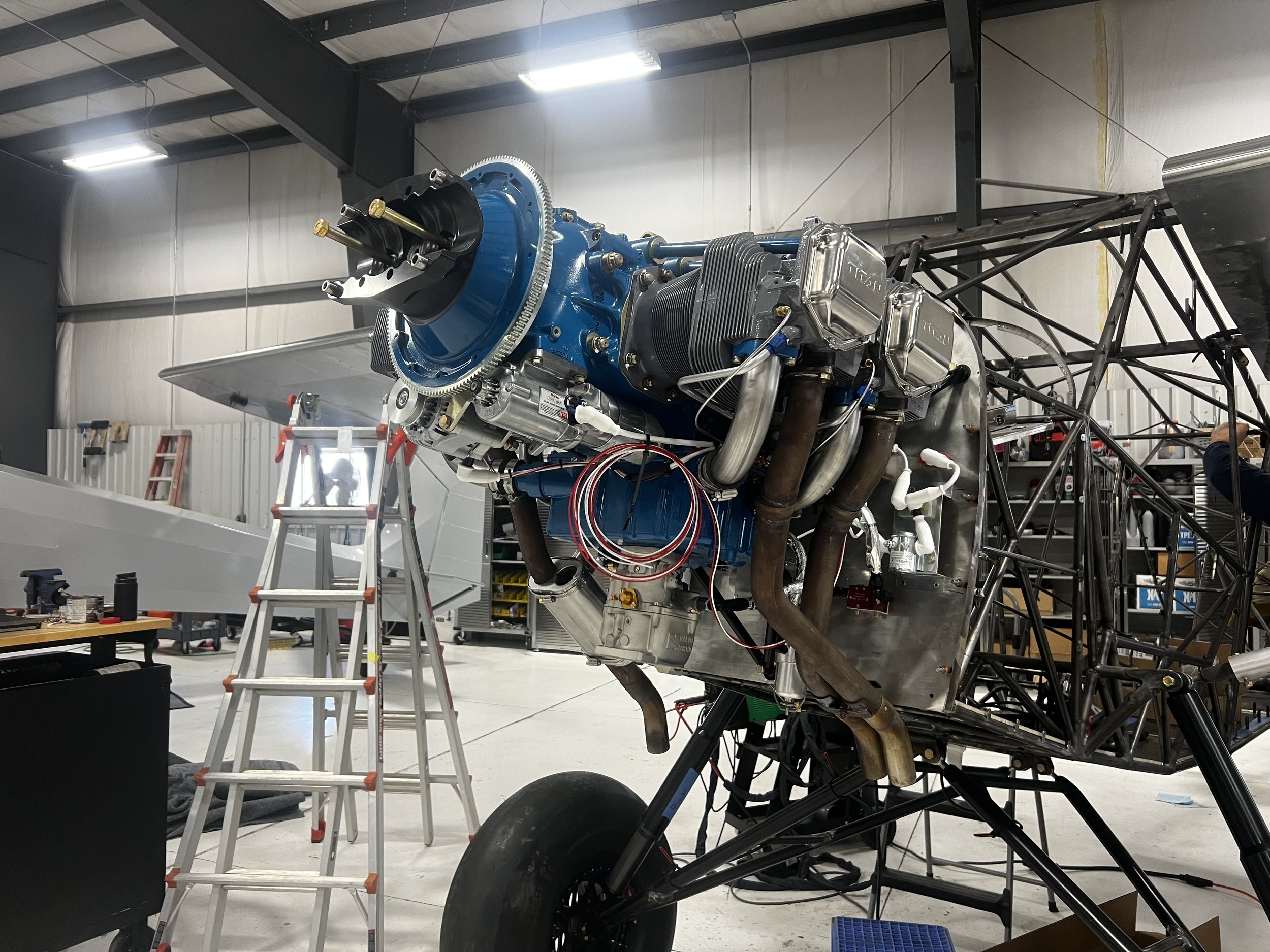 A partially assembled aircraft engine mounted on a frame inside a workshop, surrounded by ladders, tools, and equipment.