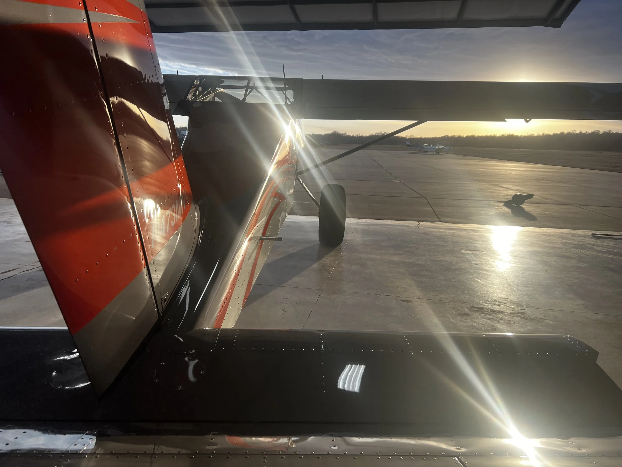 A small aircraft parked on an airport tarmac during sunset, with sunlight reflecting off the plane's shiny red, black, and silver exterior.