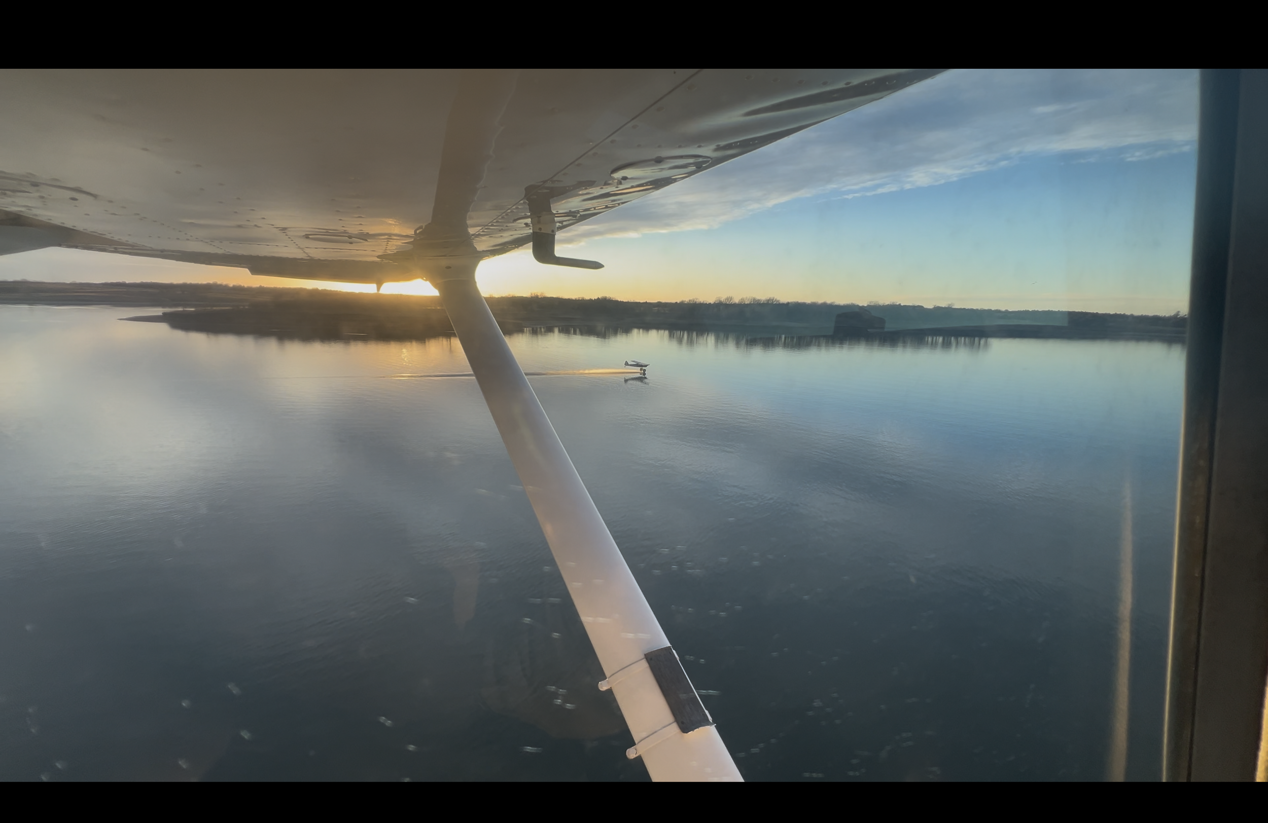 View from a small aircraft window showing a river, with a smaller plane flying close to the water at sunset or sunrise.