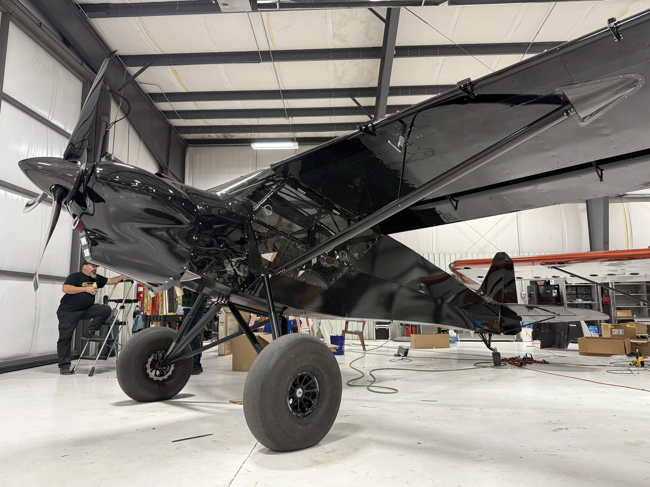 A black aircraft inside a hangar, with a person working on it, surrounded by tools and boxes.