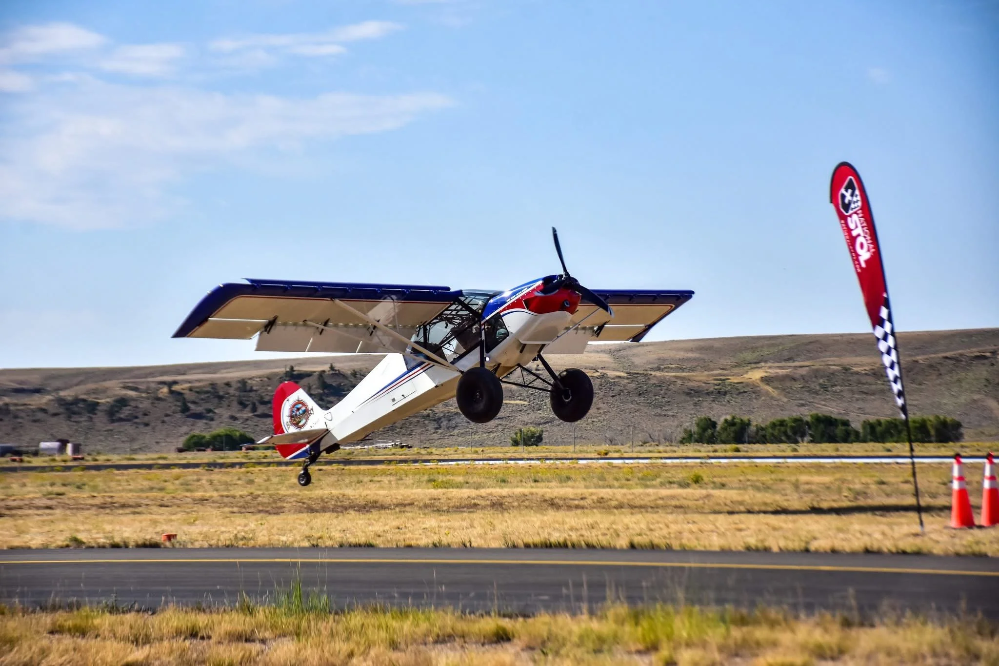 Small aircraft taking off from a runway with a red, white, and blue design, and a flag with a checkered pattern on a pole nearby.