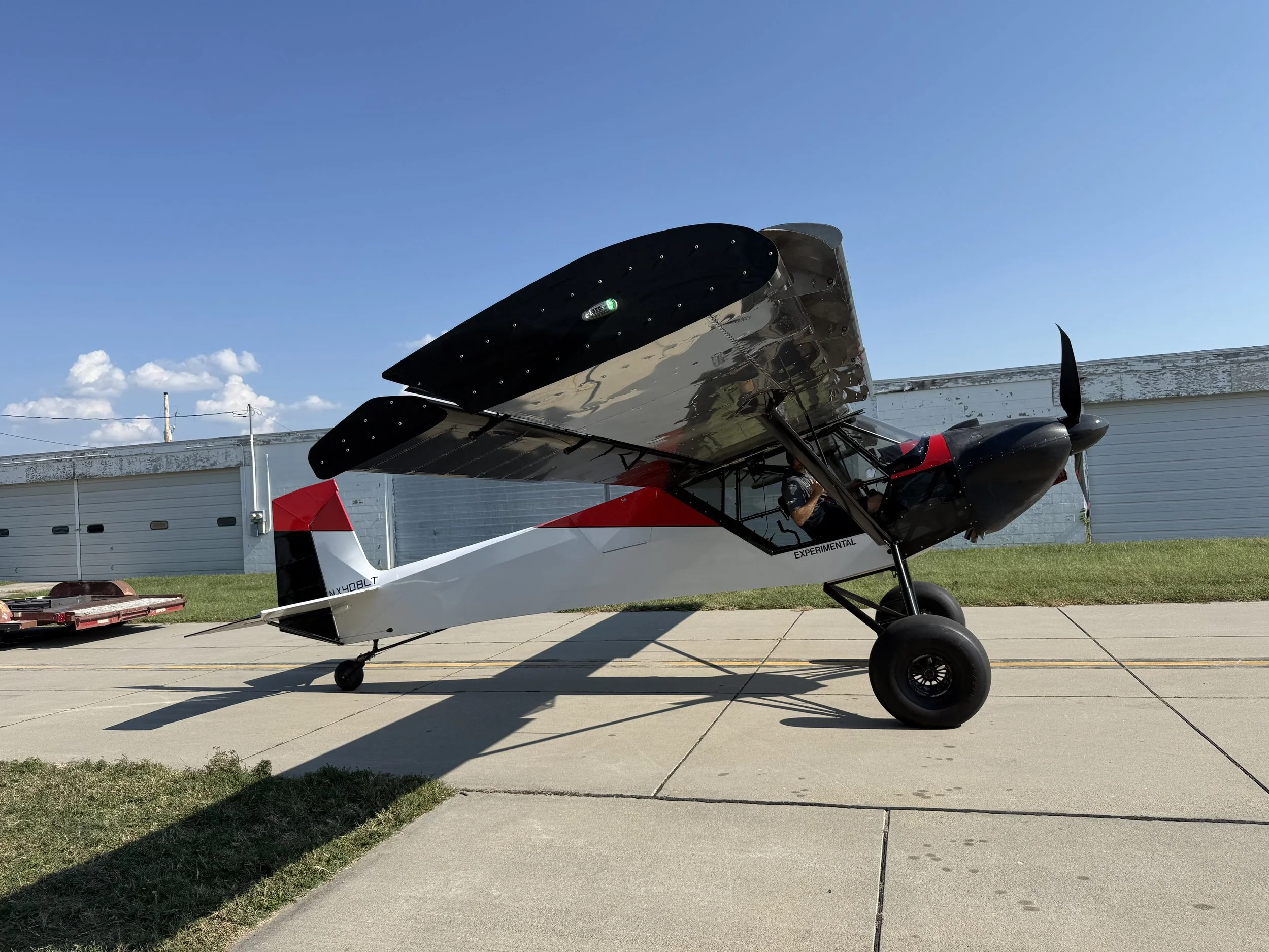 Small experimental aircraft with red and black accents parked on a concrete runway, with a blue sky and hangar in the background.