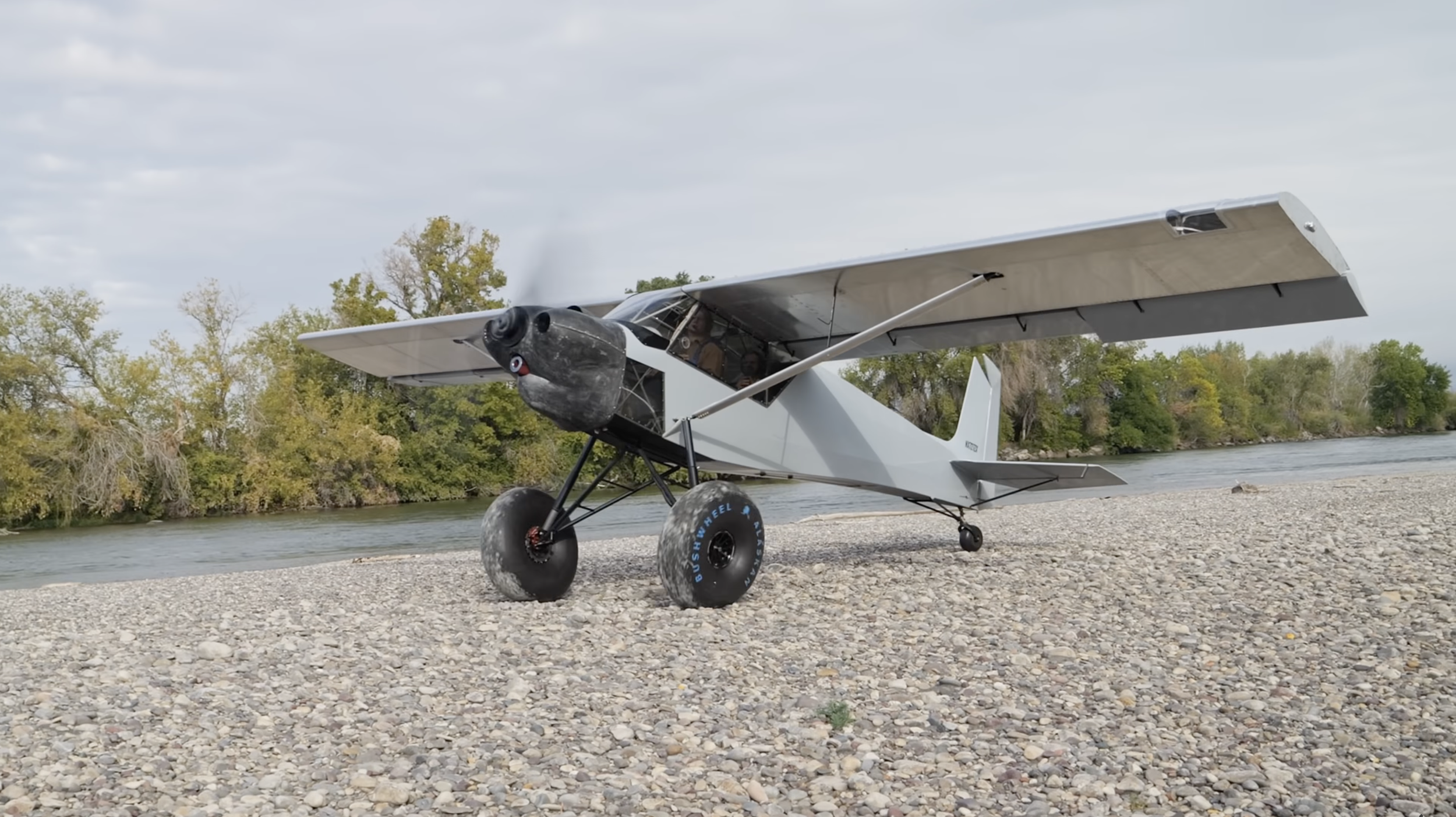 A small aircraft with large bushwheel tires parked on a rocky beach near a river, with trees in the background under a cloudy sky.