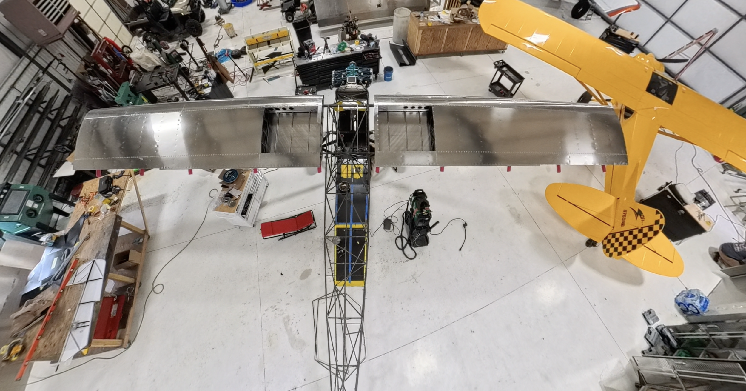 An aerial view of an aircraft under construction inside a workshop. The partially built plane has metallic wings and a black and yellow fuselage, with a smaller yellow tail fin marked with a checkered pattern. Surrounding the plane are tools, workben