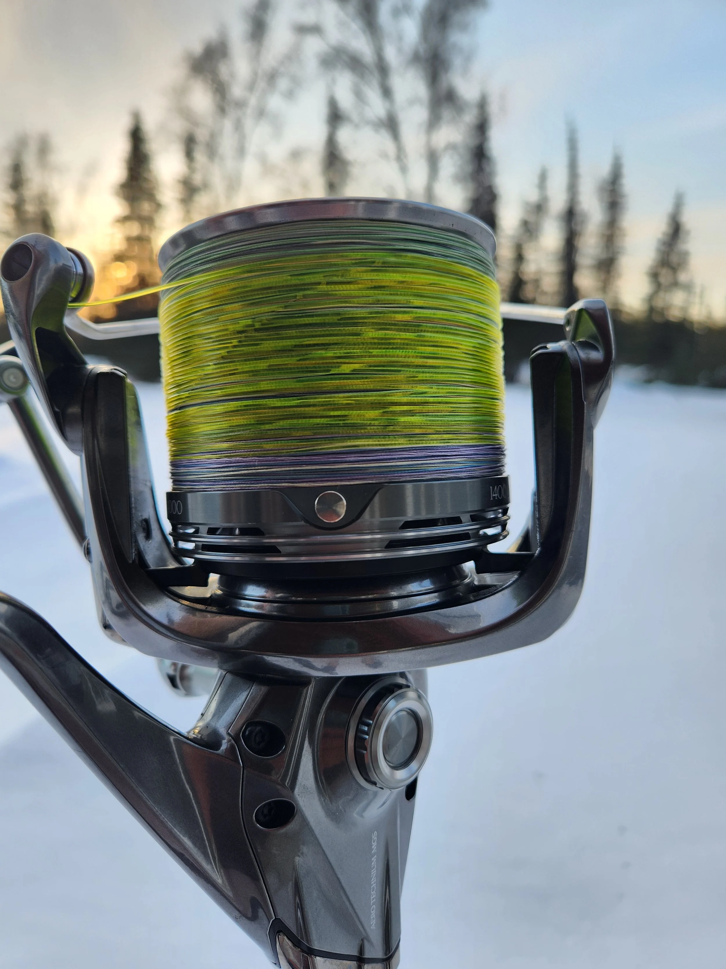 Close-up of a fishing reel with green and purple fishing line, set against a snowy outdoor landscape with trees and a sunset.