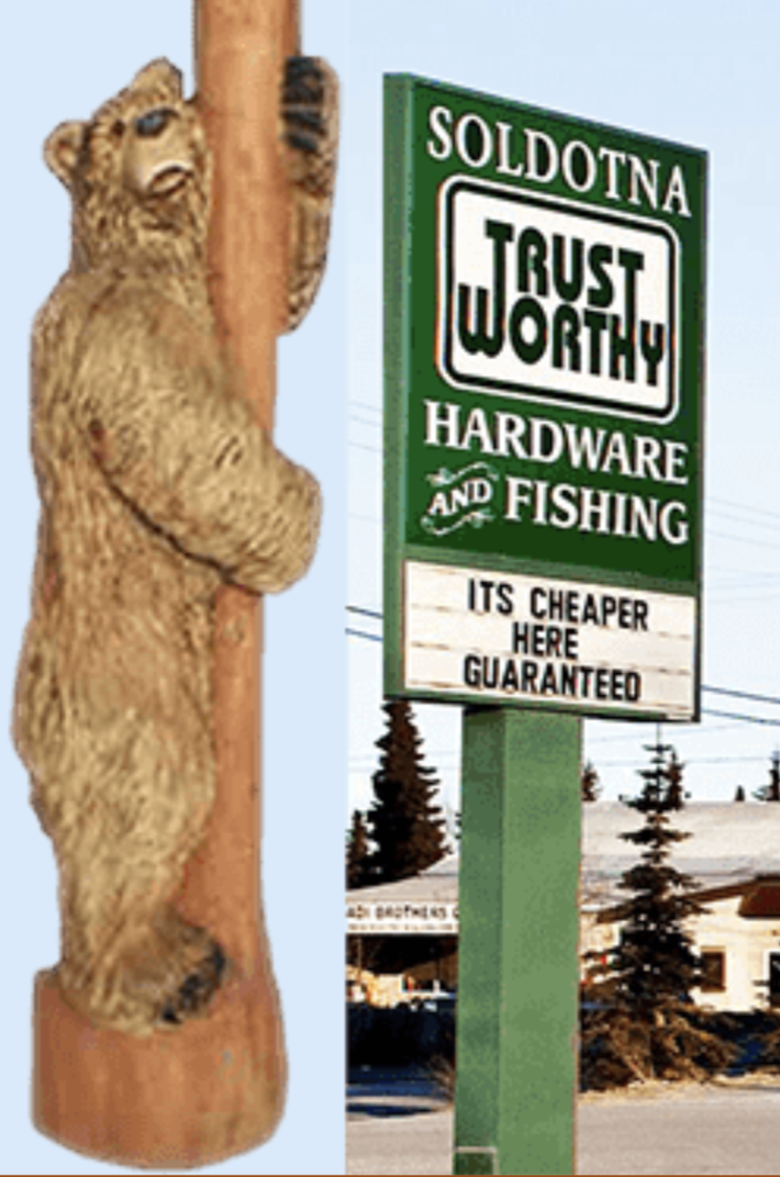 A teddy bear climbing a tree next to a sign for hardware and fishing store with a message promoting cheaper prices.