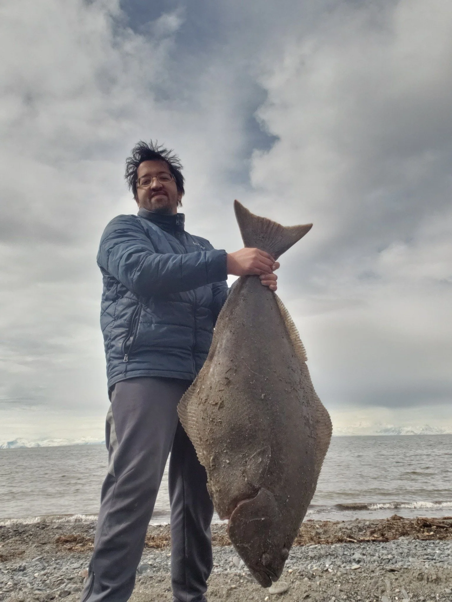 Man holding a large flatfish on a beach with cloudy sky in the background.